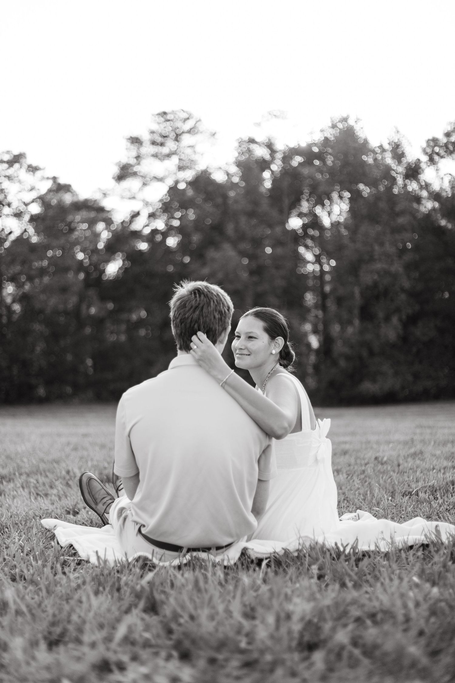 Black and white photo of couple sitting on grass in a field, woman smiling and resting arm on mans shoulder.