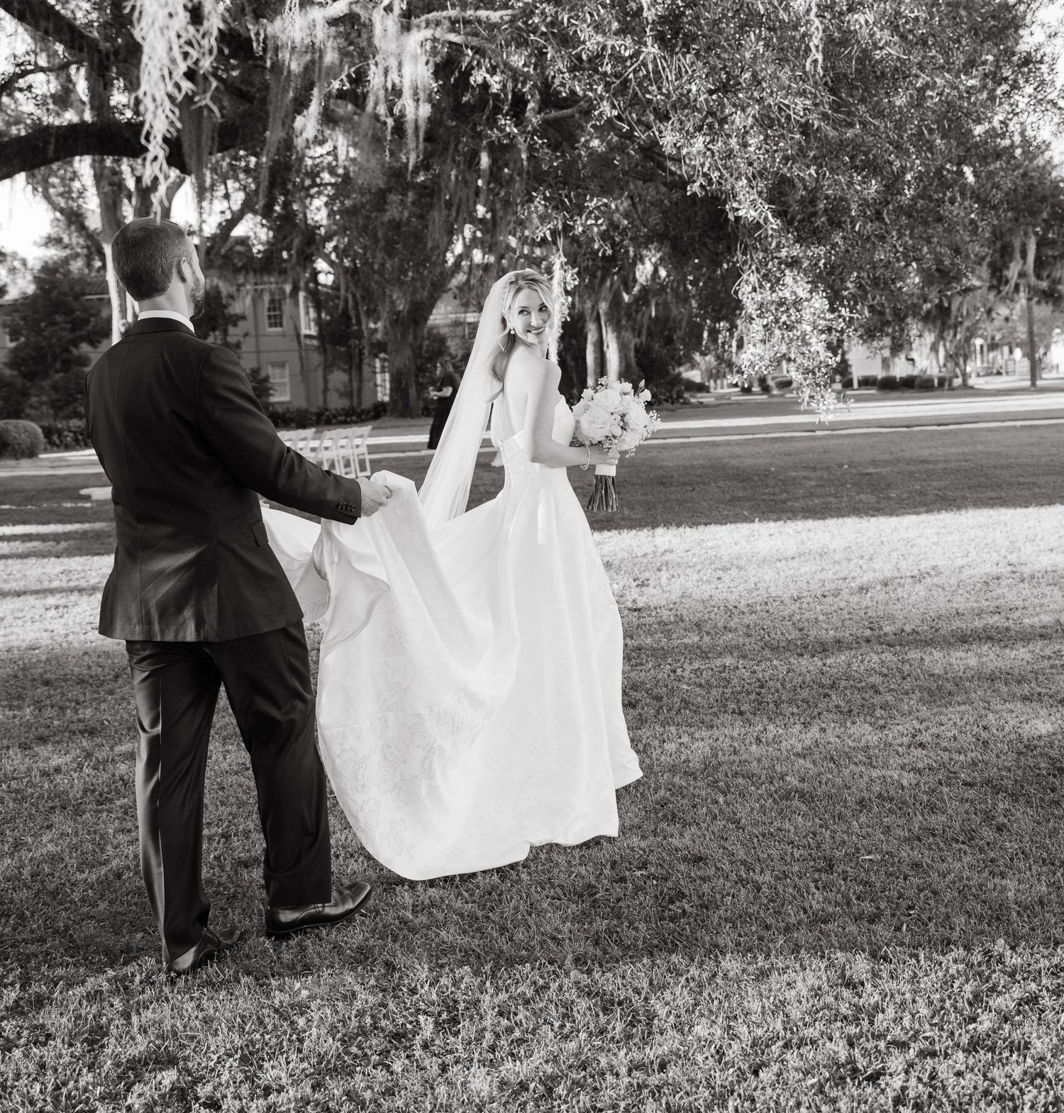 Black and white photo of a groom helping his bride with her gown train under a large oak tree.