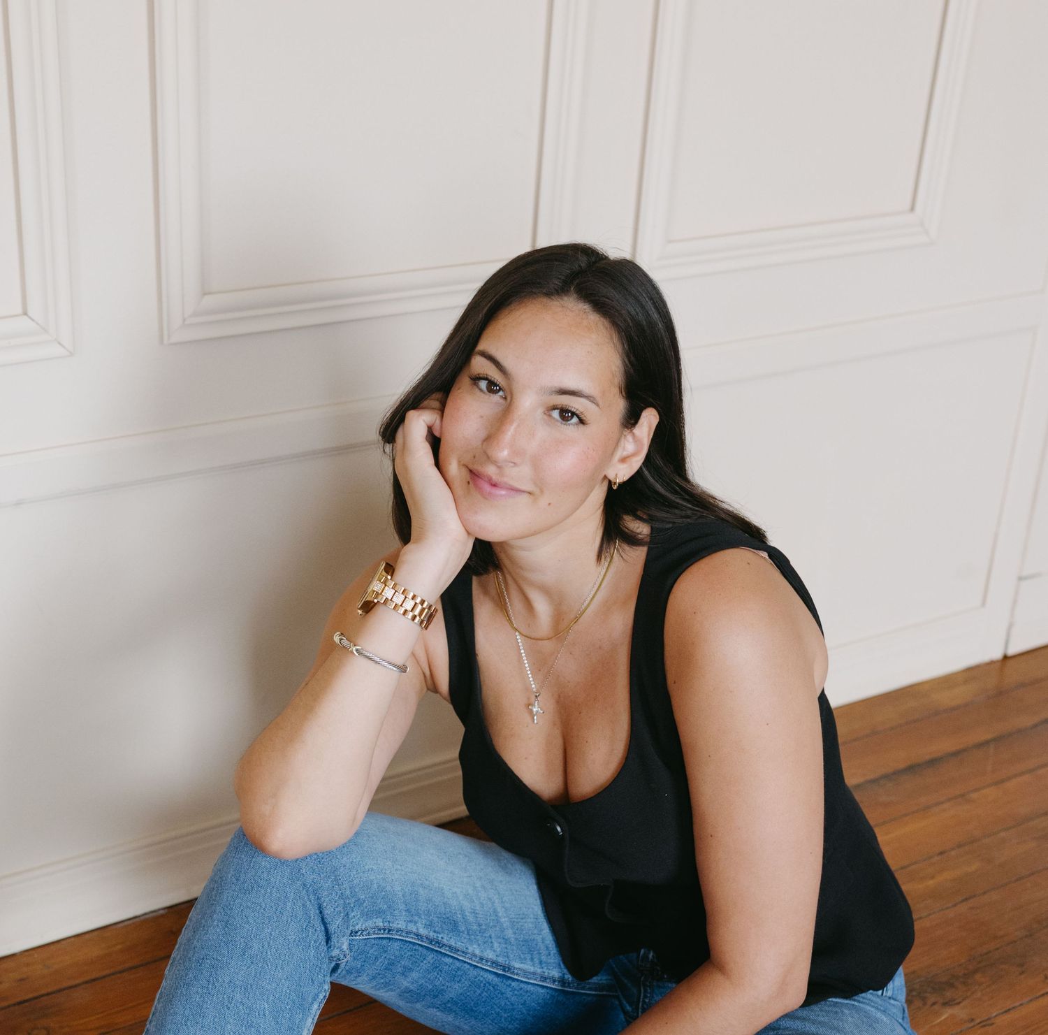 Woman in black tank top and jeans sitting on hardwood floor against white paneled wall.
