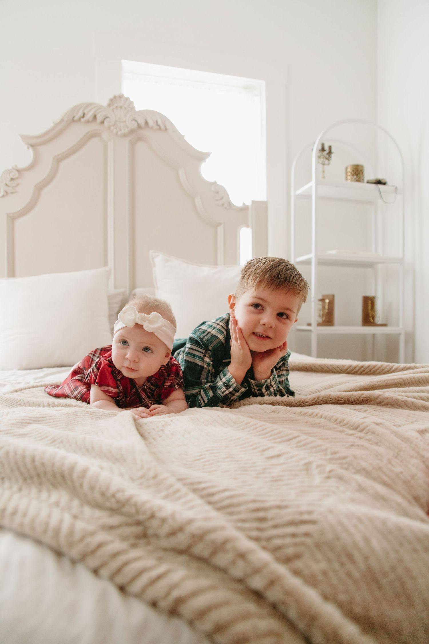 A young boy in green plaid and baby girl in red plaid lie together on a beige bedspread in a bright white bedroom.