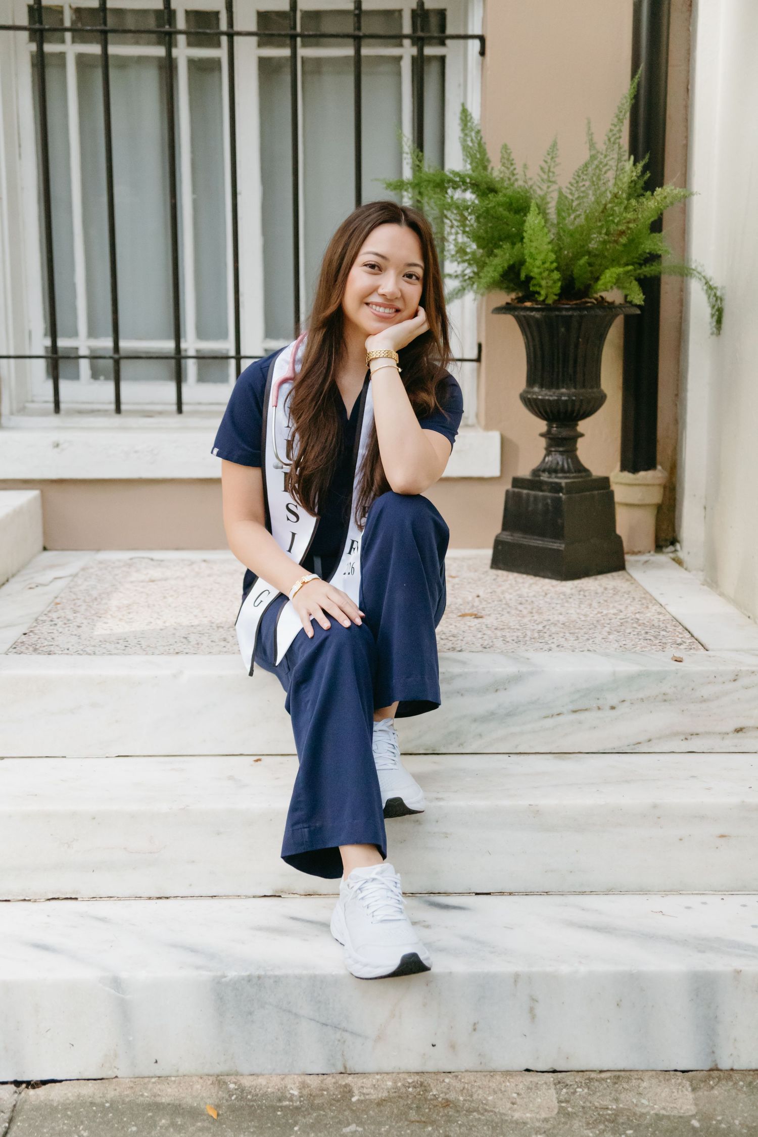 Smiling nursing graduate in scrubs and sash sitting on marble steps outdoors.