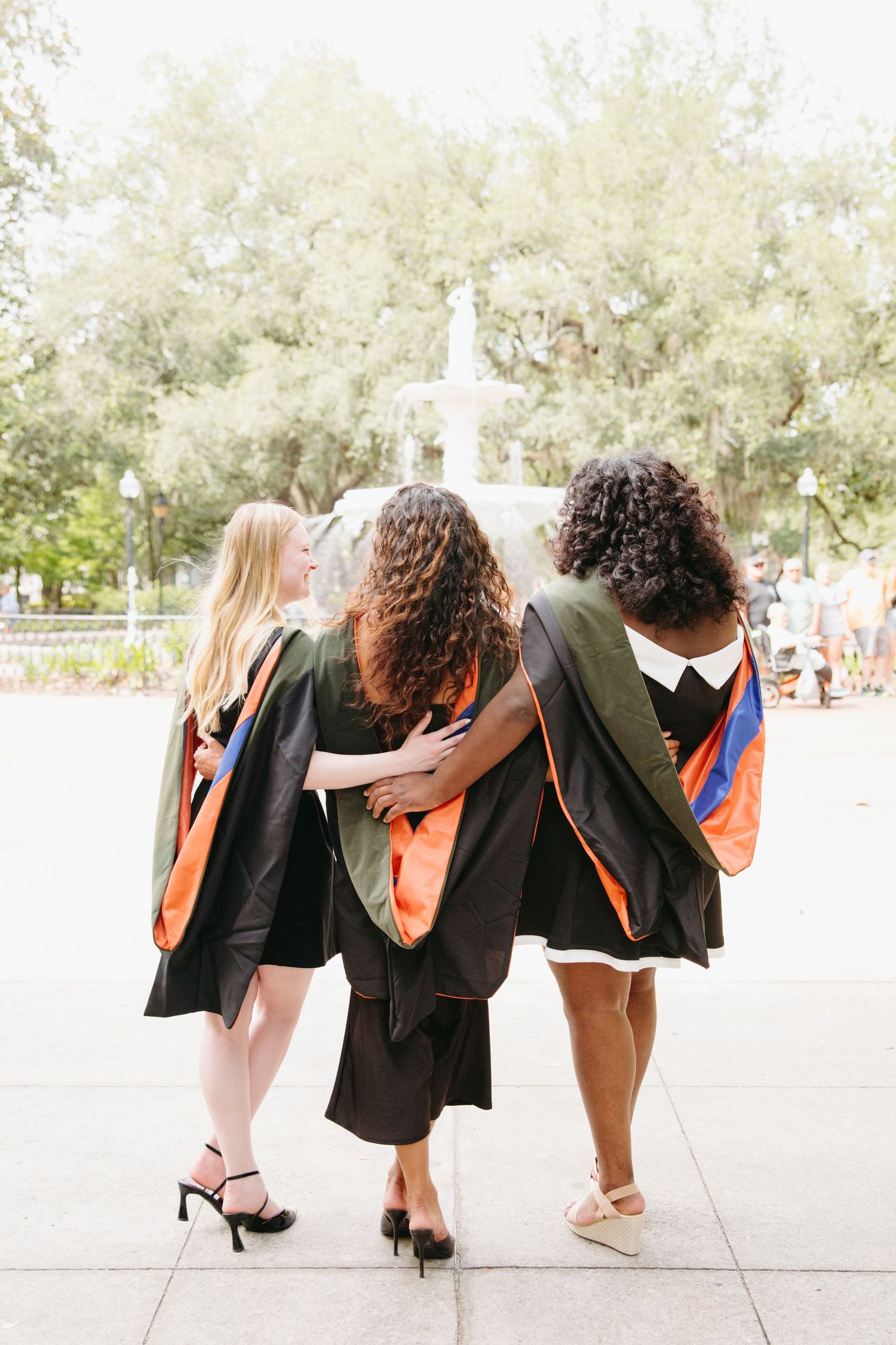 Three graduates in black gowns with orange stoles walking toward park fountain.