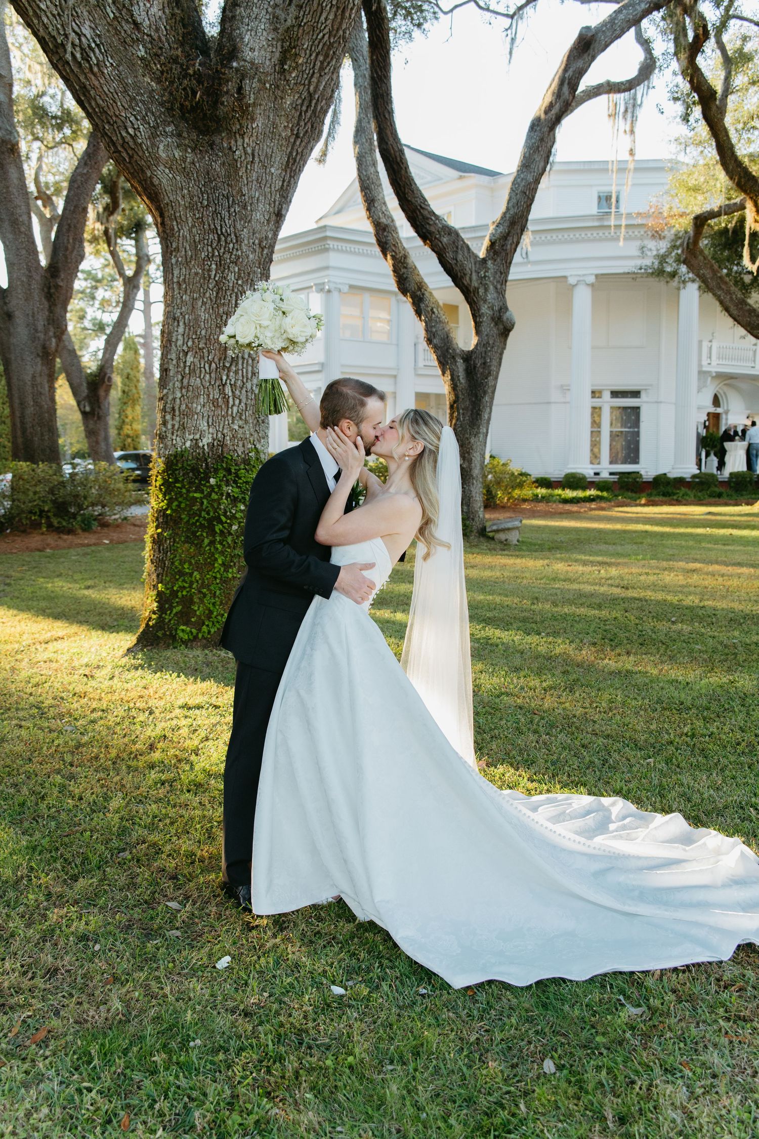 Bride and groom sharing a kiss outdoors near large oak trees in front of a white plantation home.