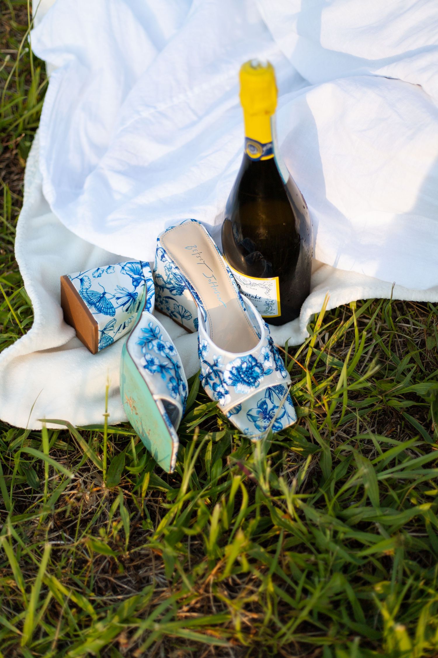 Blue floral bridal heels and champagne bottle resting on white fabric in green grass.
