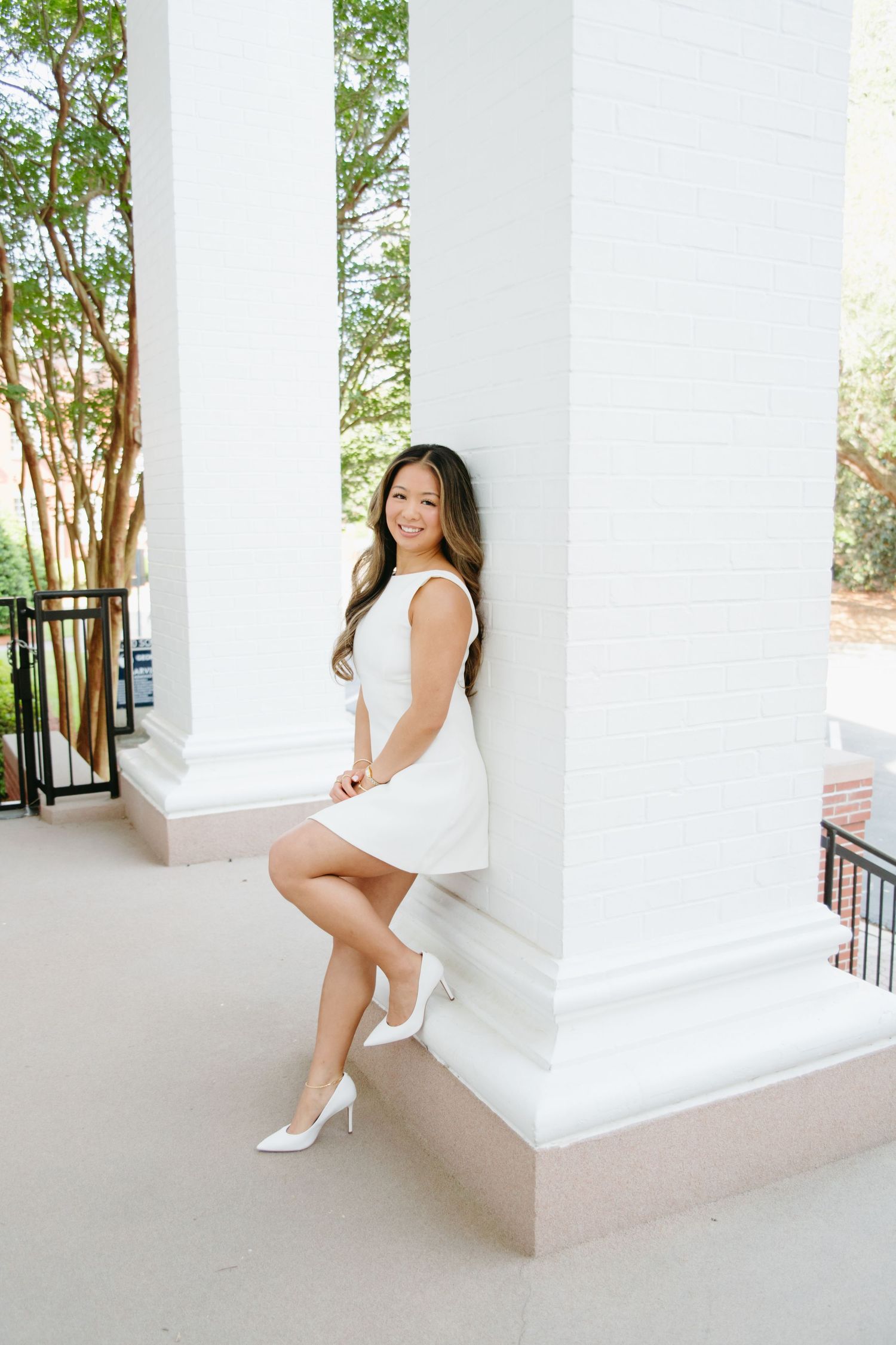 Young woman in white mini dress and heels leaning against white column on elegant porch for graduation photos.