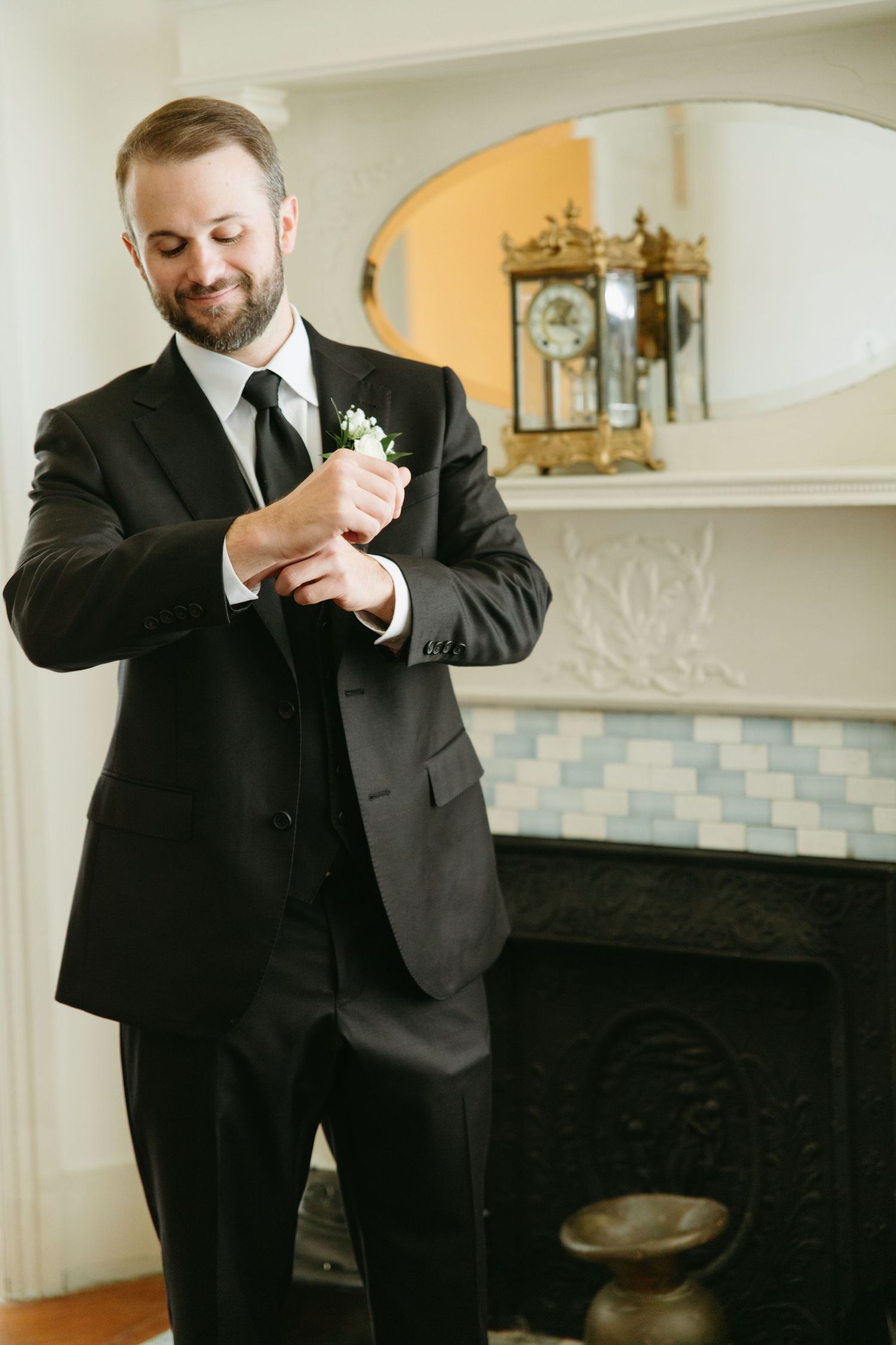 Dapper groom in black suit adjusts cufflinks before fireplace on his wedding day.