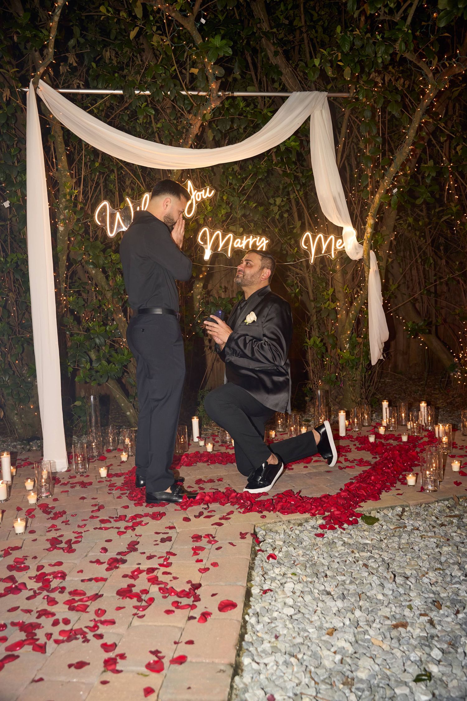 A romantic same-sex marriage proposal with neon sign, rose petals, and candles at night.