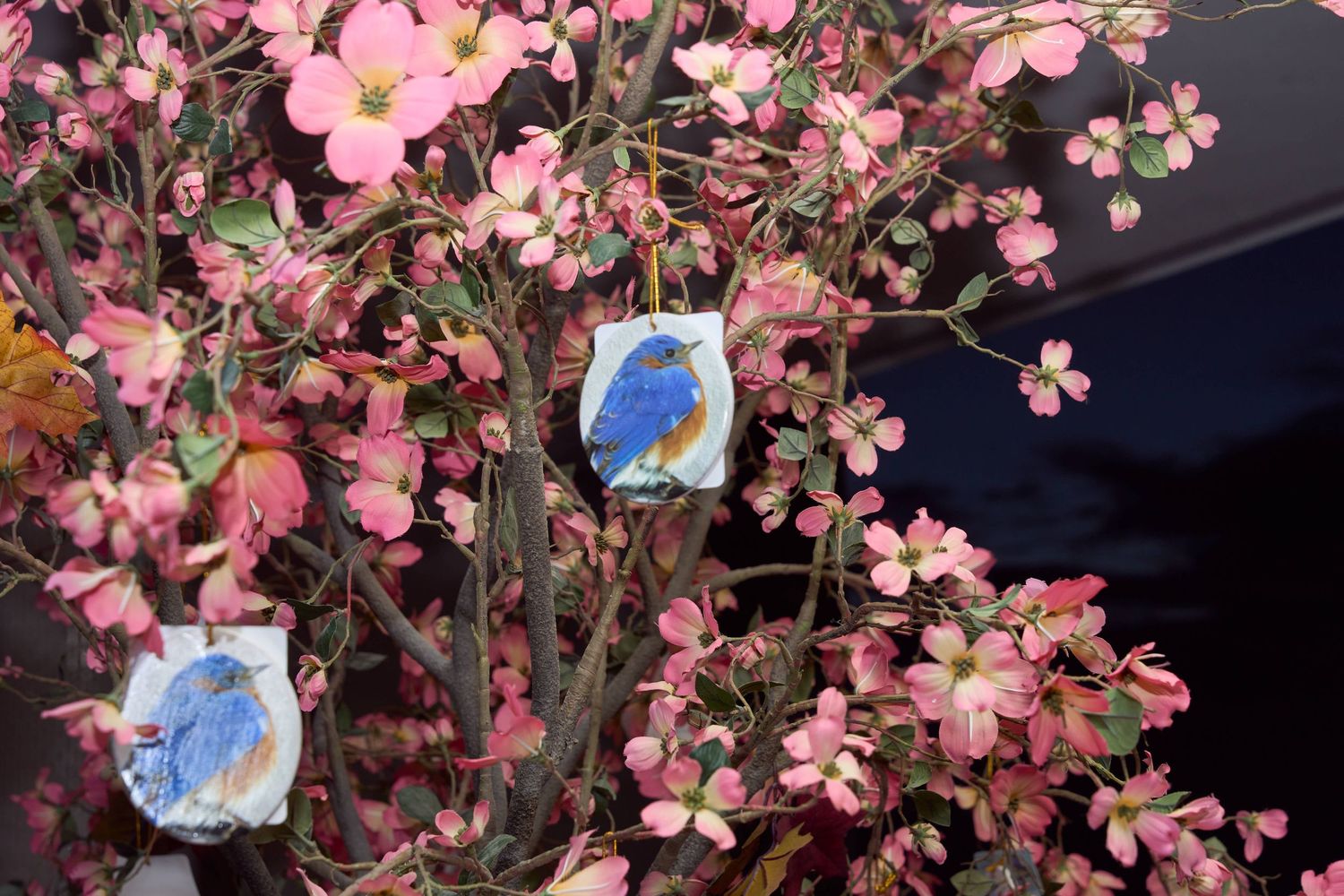 Two bluebird ornaments hang among pink dogwood blossoms on a flowering tree branch in spring.