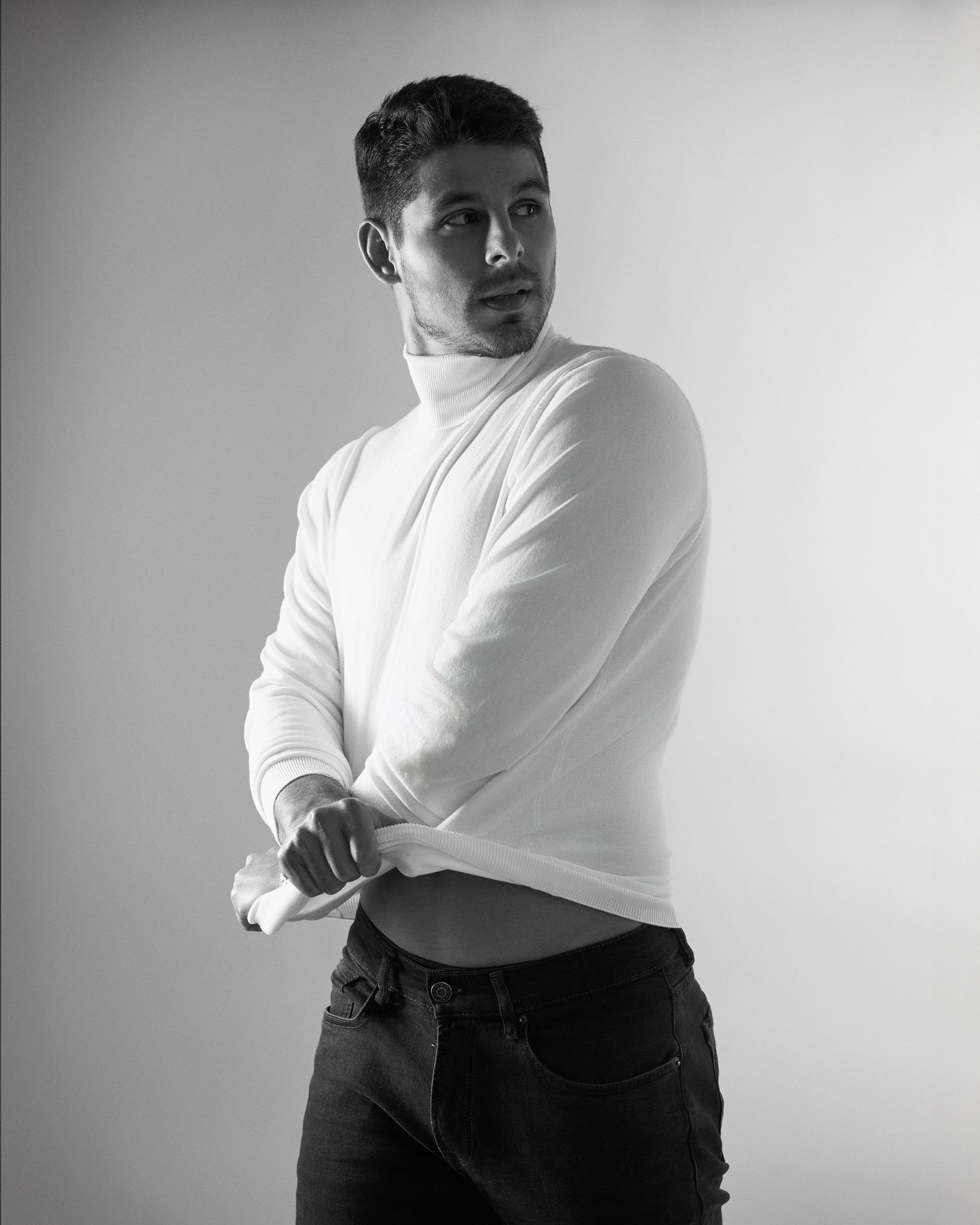Black and white portrait of a young man in a white turtleneck sweater and dark jeans posing confidently.