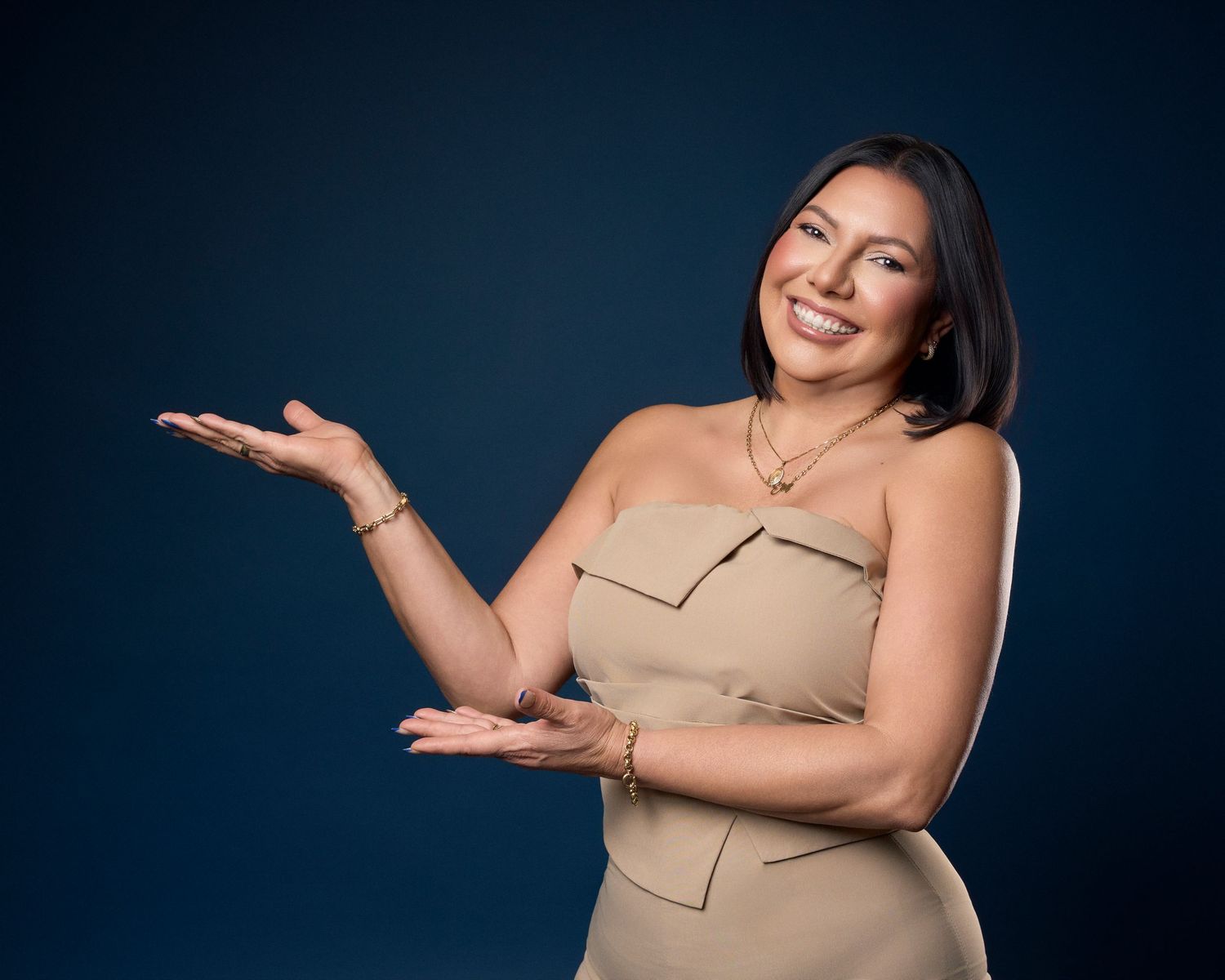 Smiling woman in beige strapless dress gesturing with open hands against dark blue background.