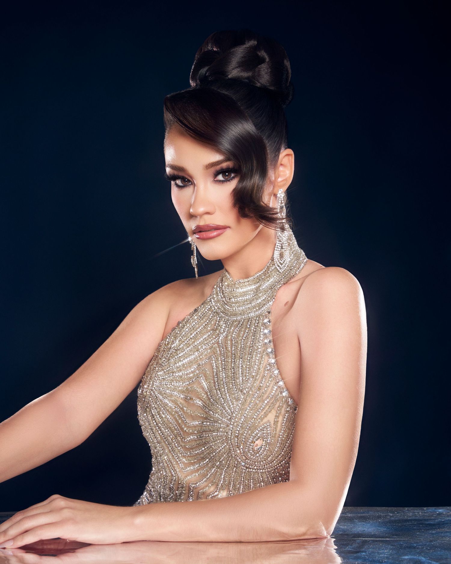 Woman in gold beaded halter gown posing at table in dark setting.