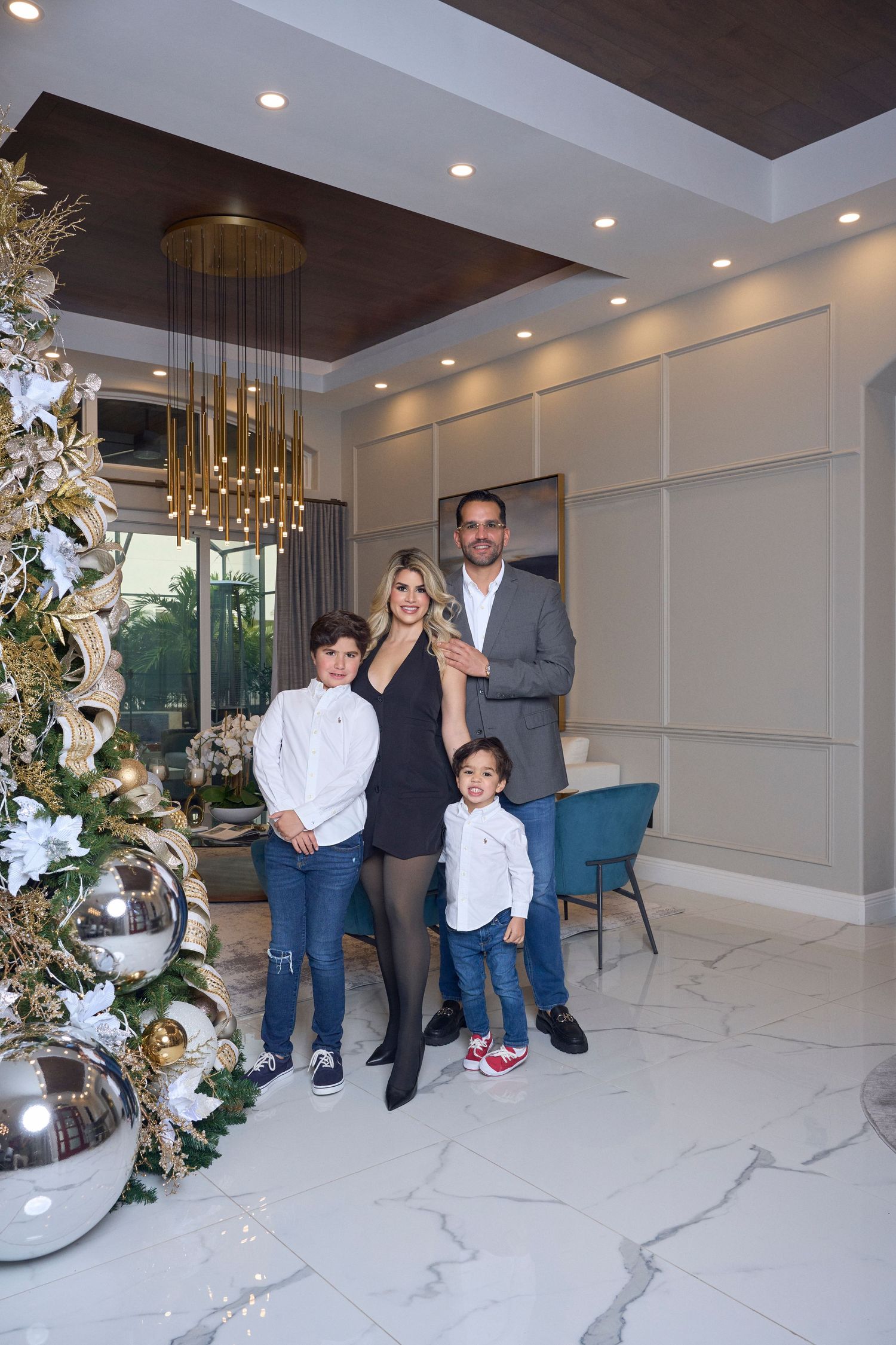 Family of four stands by ornate Christmas tree in elegant marble-floored home.