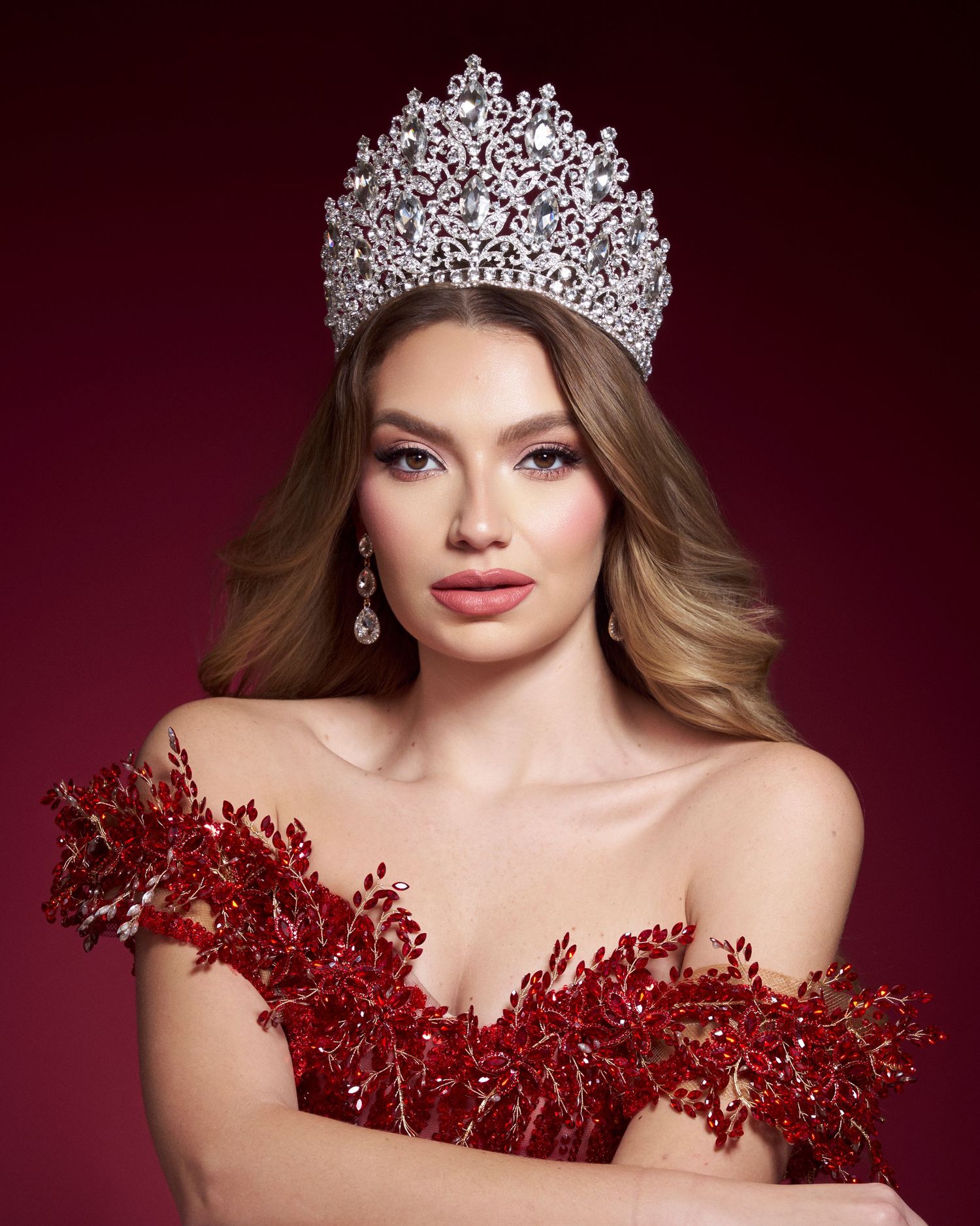 Beauty queen in red feathered gown wearing a large silver rhinestone crown with wavy blonde hair.