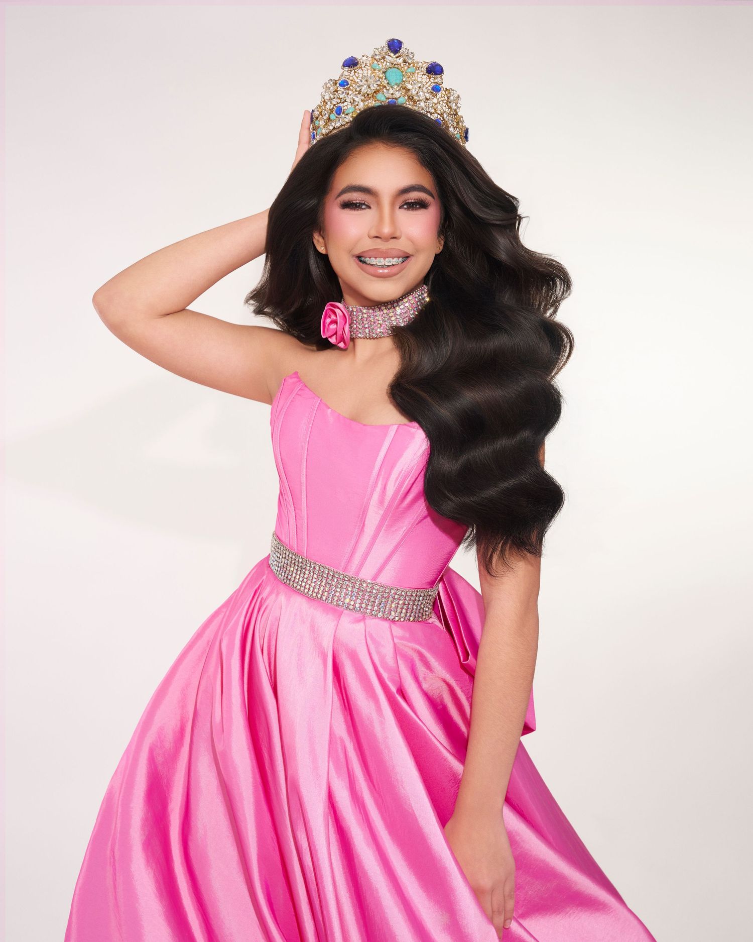 Smiling woman in pink satin gown wearing jeweled crown and rose choker.