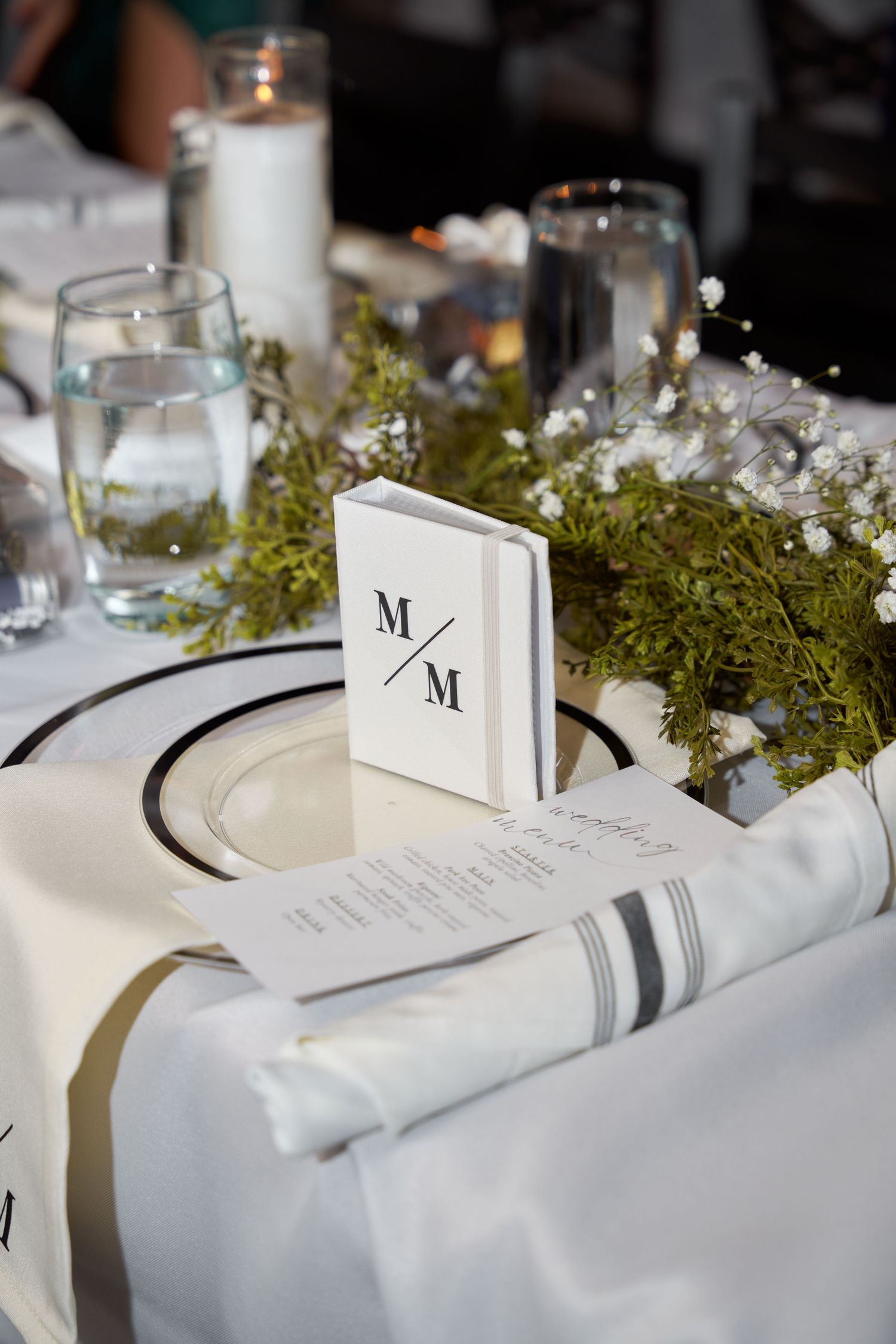 Elegant wedding table setting with monogrammed M/M card, white florals, candles, and classic black-rimmed china plates.