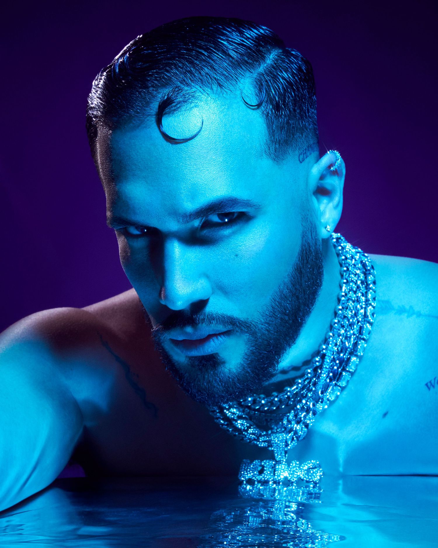 Dramatic blue-lit portrait of a man with slicked hair wearing a diamond chain necklace emerging from water.