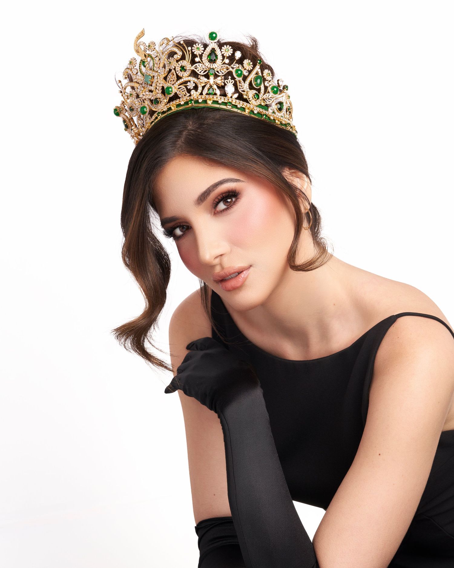 Woman in black dress and long gloves wearing gold emerald pageant crown.