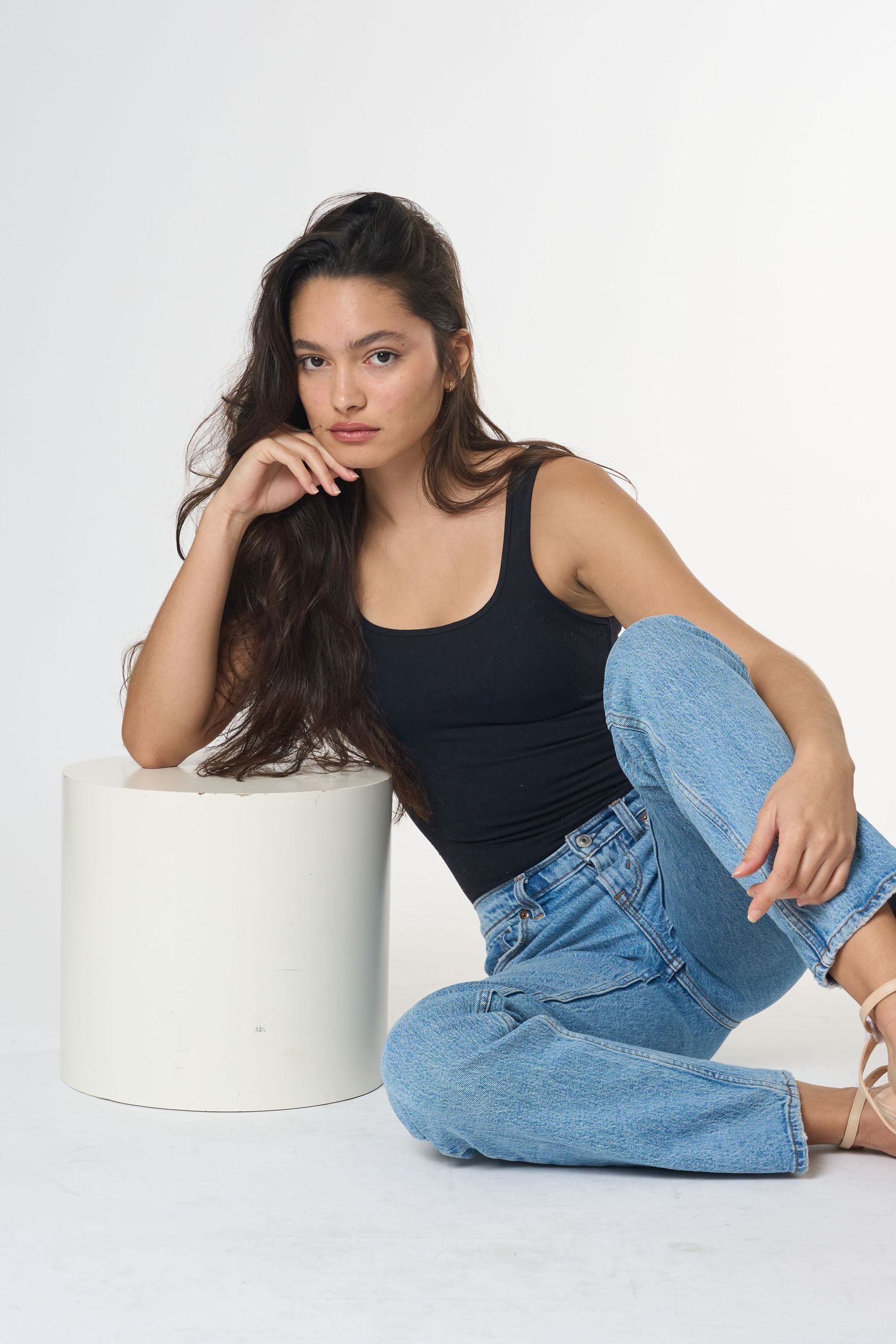Woman in black tank top and blue jeans sitting on floor leaning against white cylinder prop.