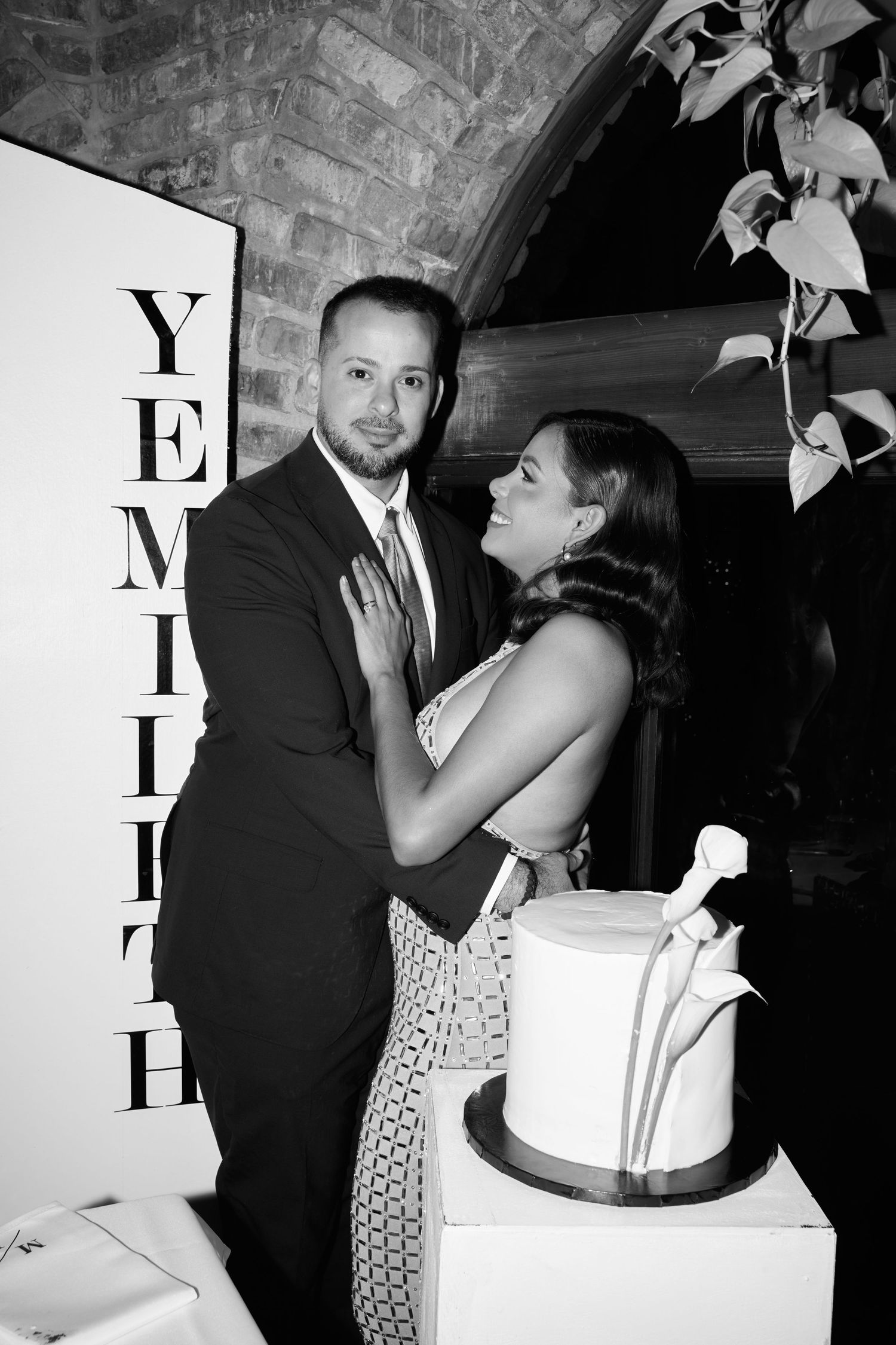 Black and white photo of a couple embracing near a wedding cake at an elegant venue with brick arched ceiling.