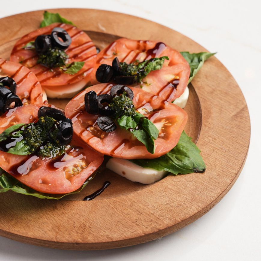 Caprese salad with tomatoes, mozzarella, basil, black olives, and balsamic glaze on wood board.