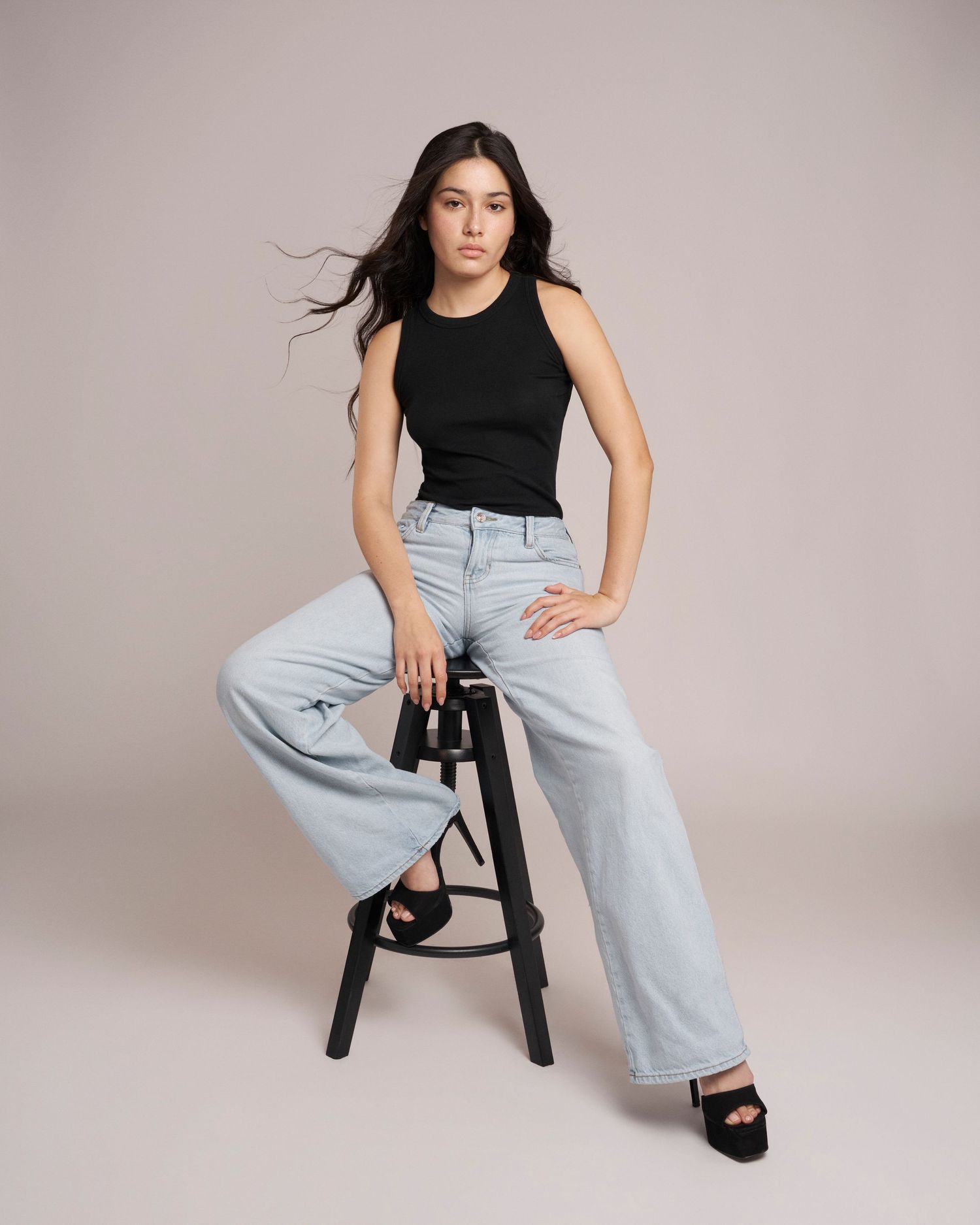 Young woman in black tank top and wide-leg light jeans posing on stool against beige background.