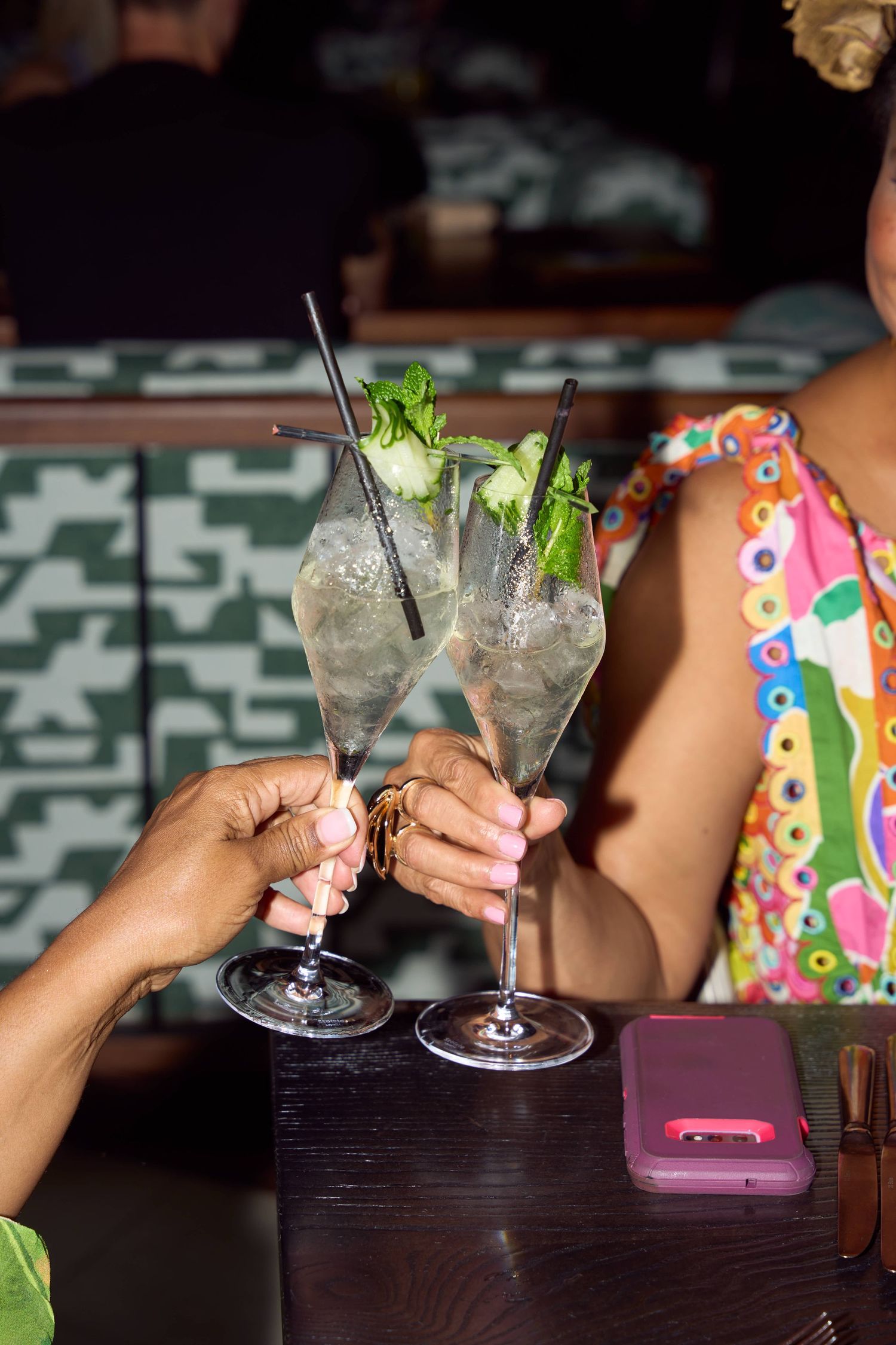 Two women toasting with mojito cocktails in champagne glasses at a bar, garnished with mint and black straws.