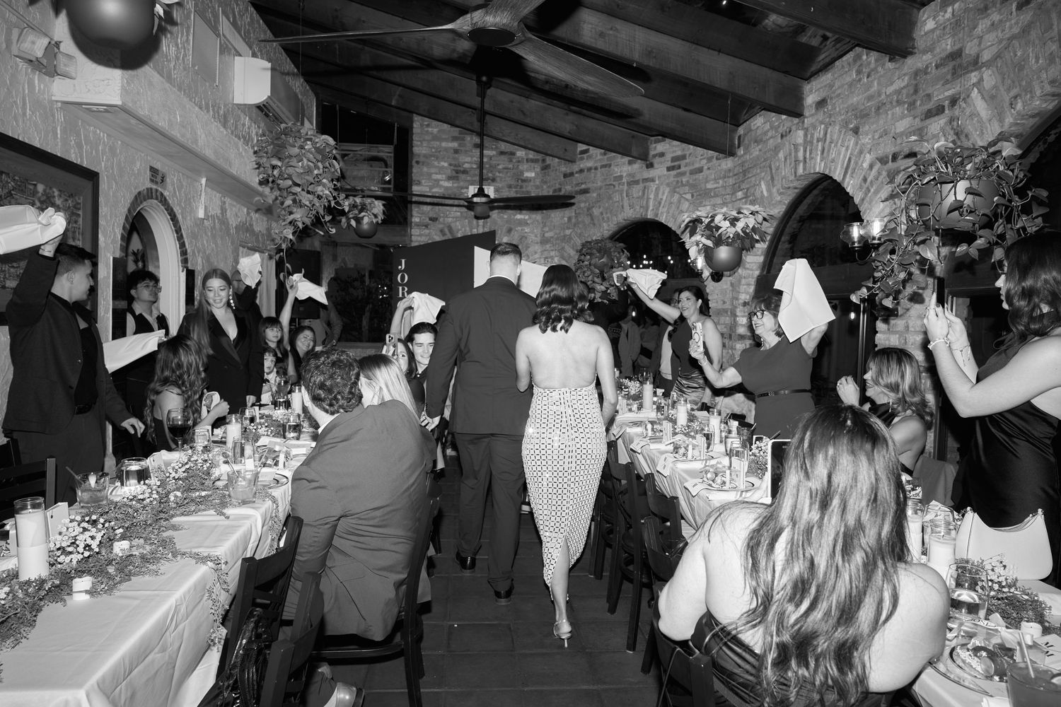 Black and white photo of elegant wedding reception dinner in rustic stone venue with guests dining at long tables.