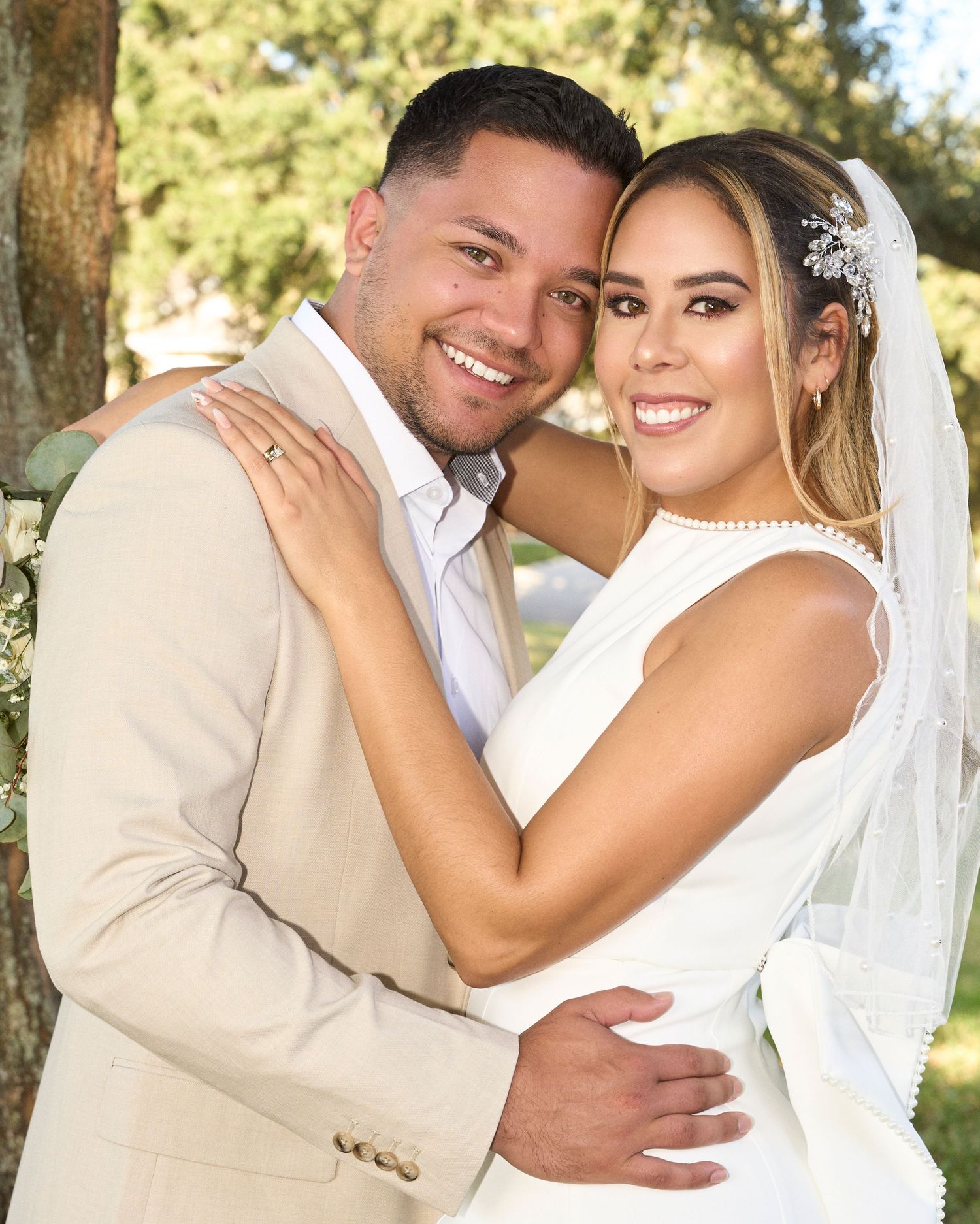 Smiling bride and groom in tan suit embrace warmly during outdoor wedding portrait under trees.