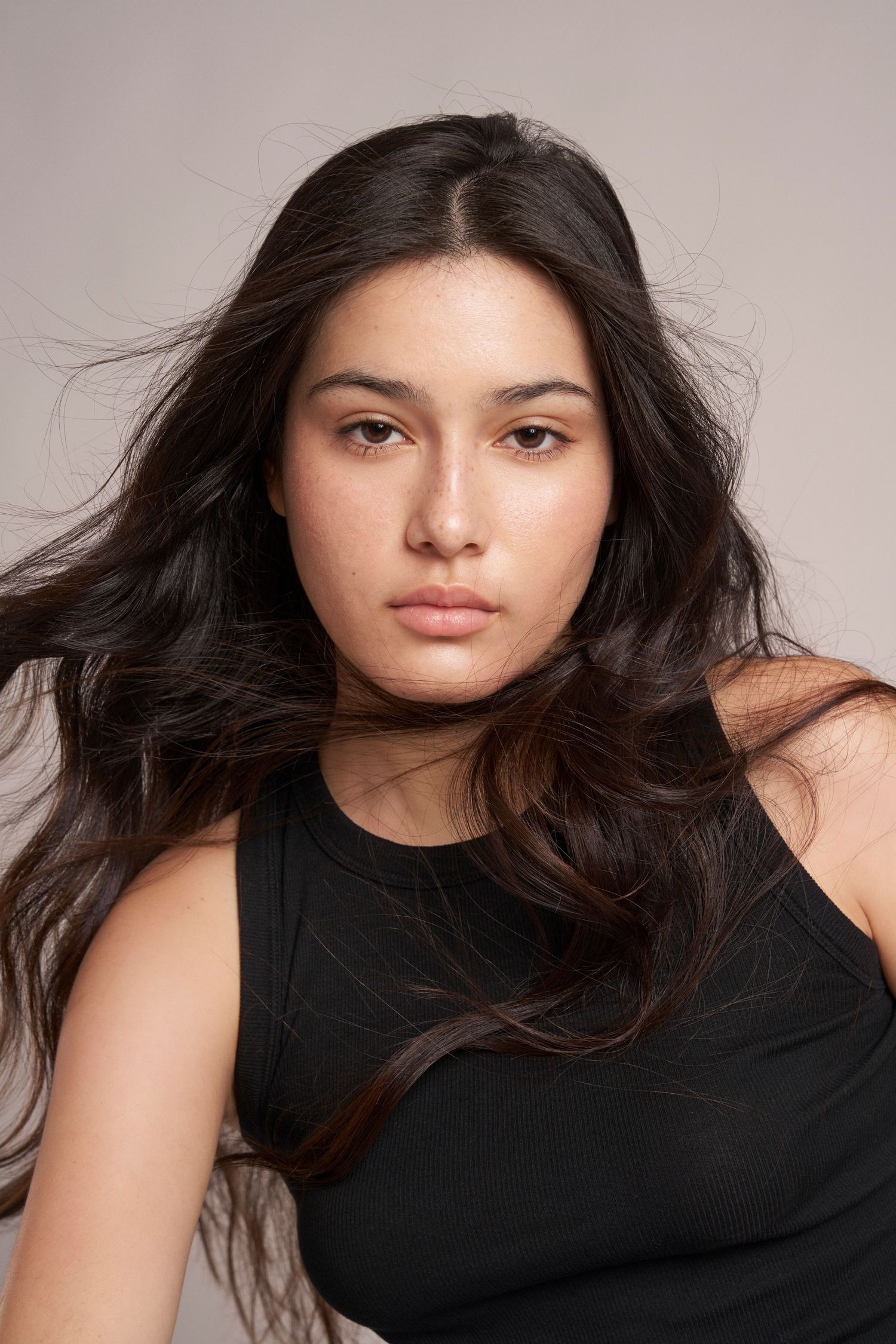 Close-up portrait of young woman with long dark flowing hair in black top on gray background.