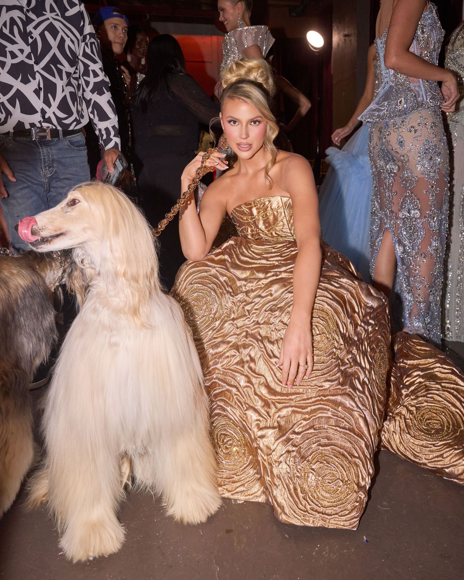 Blonde model in gold strapless ball gown poses with Afghan Hound at fashion event.