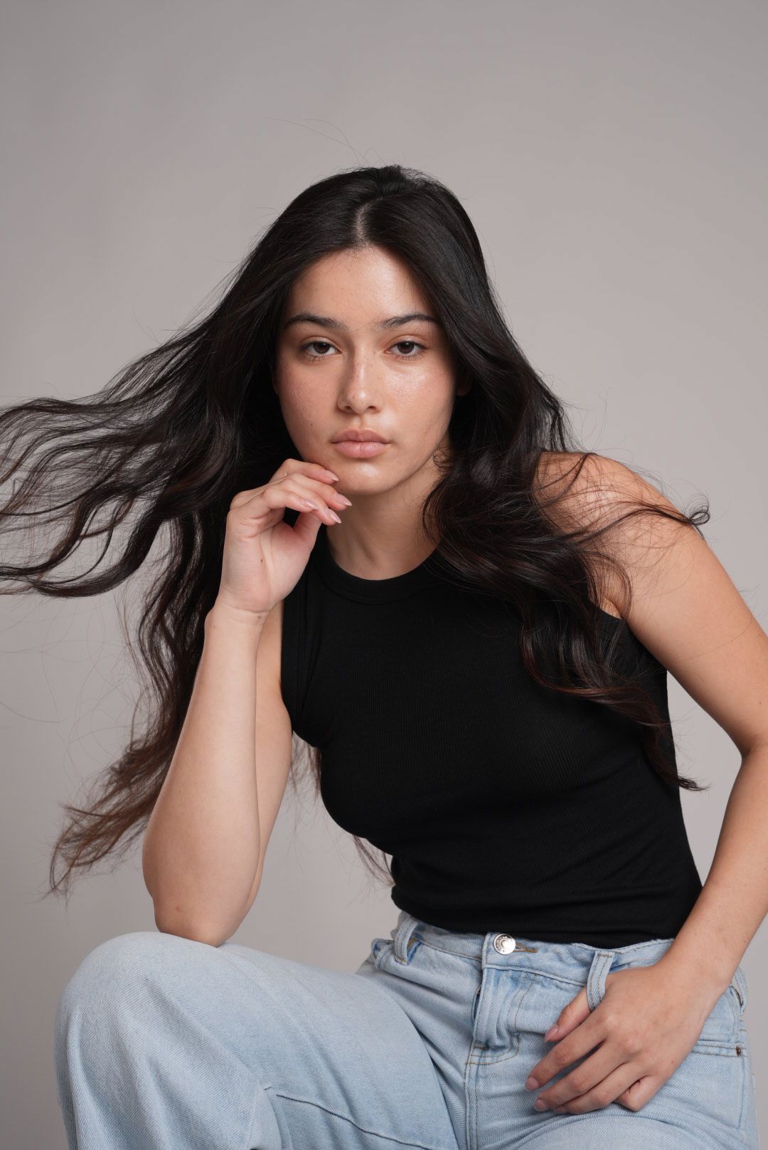 Young woman with long flowing dark hair wearing a black tank top and jeans posing in a studio portrait.