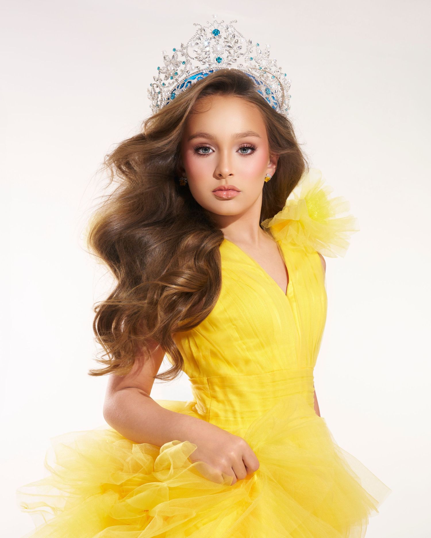 Young girl wearing a sparkling tiara and yellow ruffled gown with flowing wavy hair in a pageant portrait.