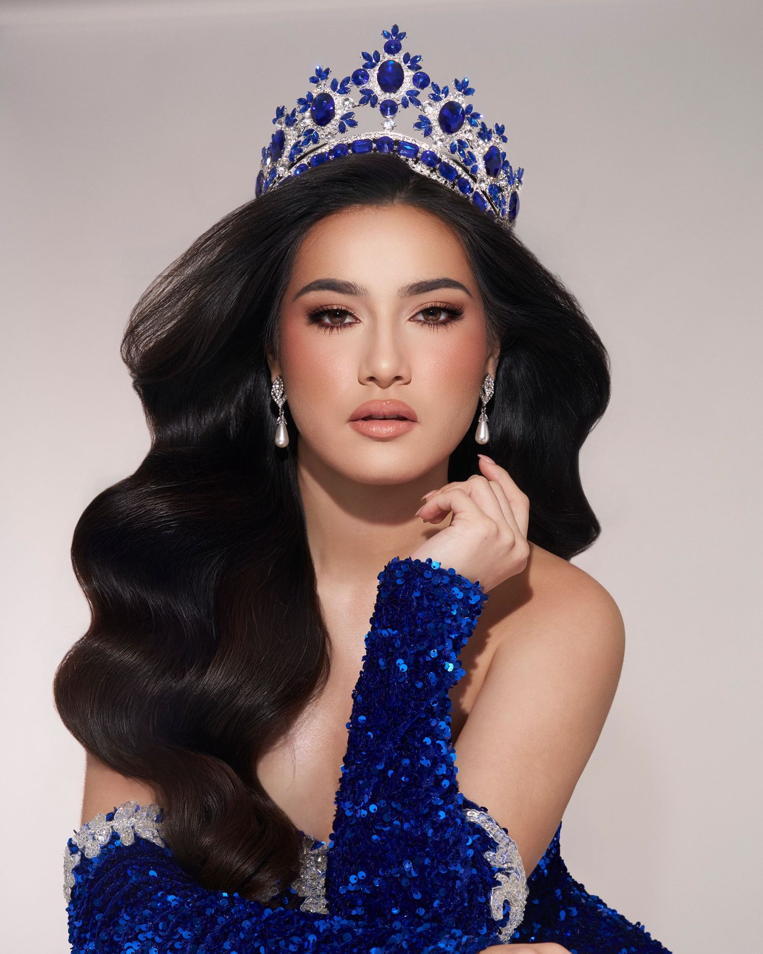 Woman wearing a blue sequin gown and ornate blue crystal crown with elegant wavy black hair.