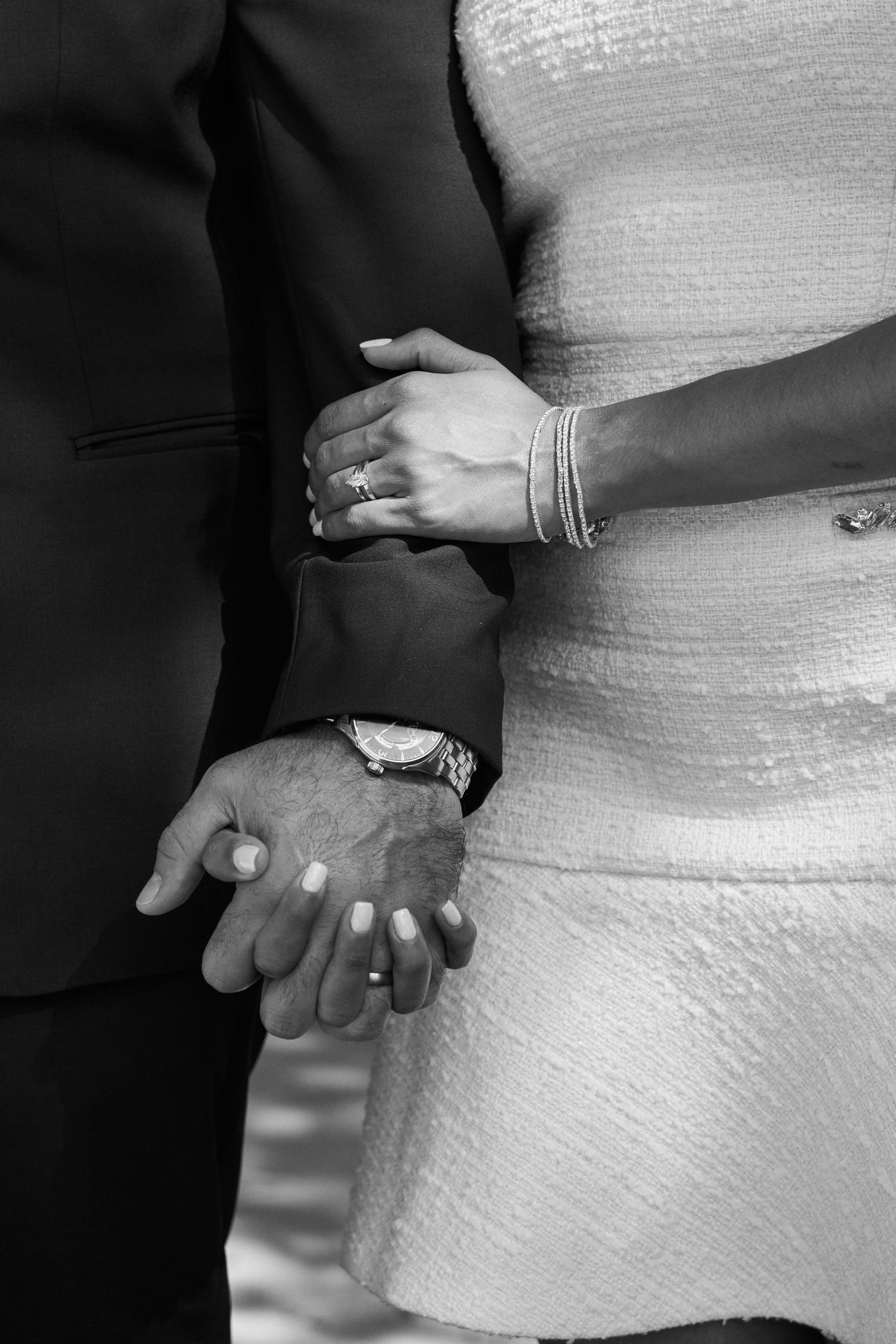 Black and white close-up of a couple holding hands, showcasing bracelets, rings, and a watch during a formal occasion.