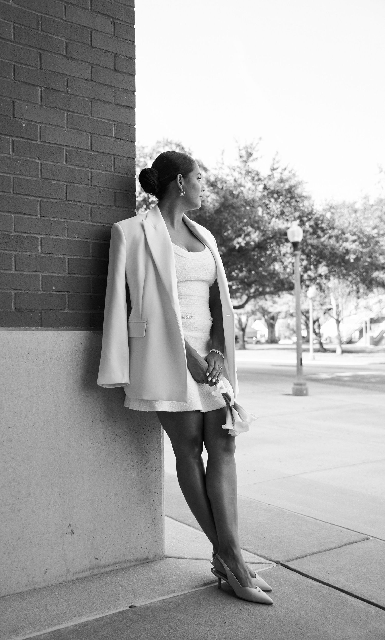 Black and white full-length portrait of a woman in a white blazer and skirt leaning against a brick column outdoors.