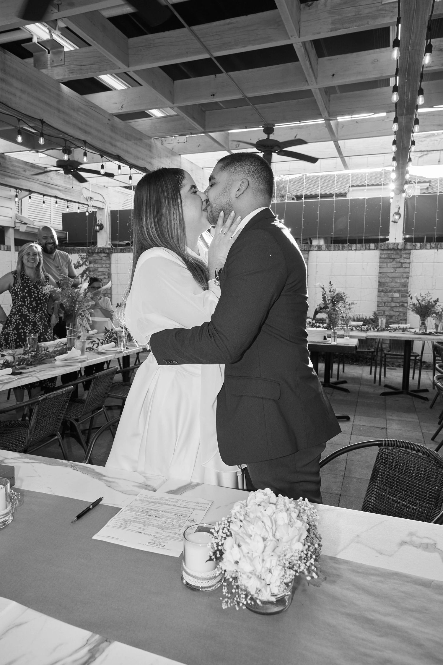 Black and white photo of a couple sharing a kiss at their wedding ceremony, with guests and floral arrangements visible.