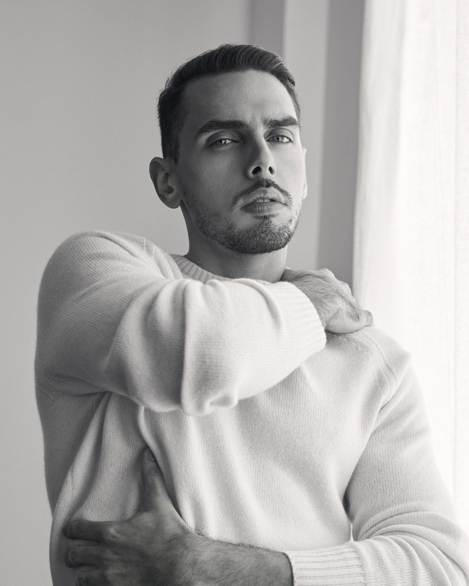 Black and white portrait of a young man in a white sweater with arms crossed, gazing thoughtfully to the side.