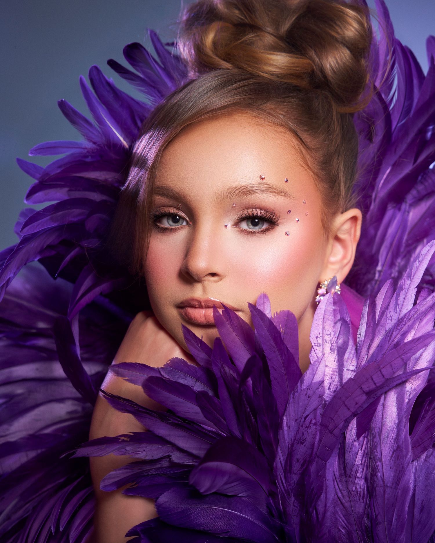 Young woman surrounded by vibrant purple feathers with elegant updo and sparkling eye makeup in studio.
