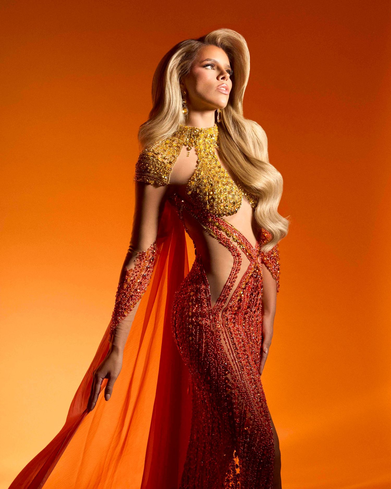 Model in gold and red beaded cutout gown with cape poses against vibrant orange background.