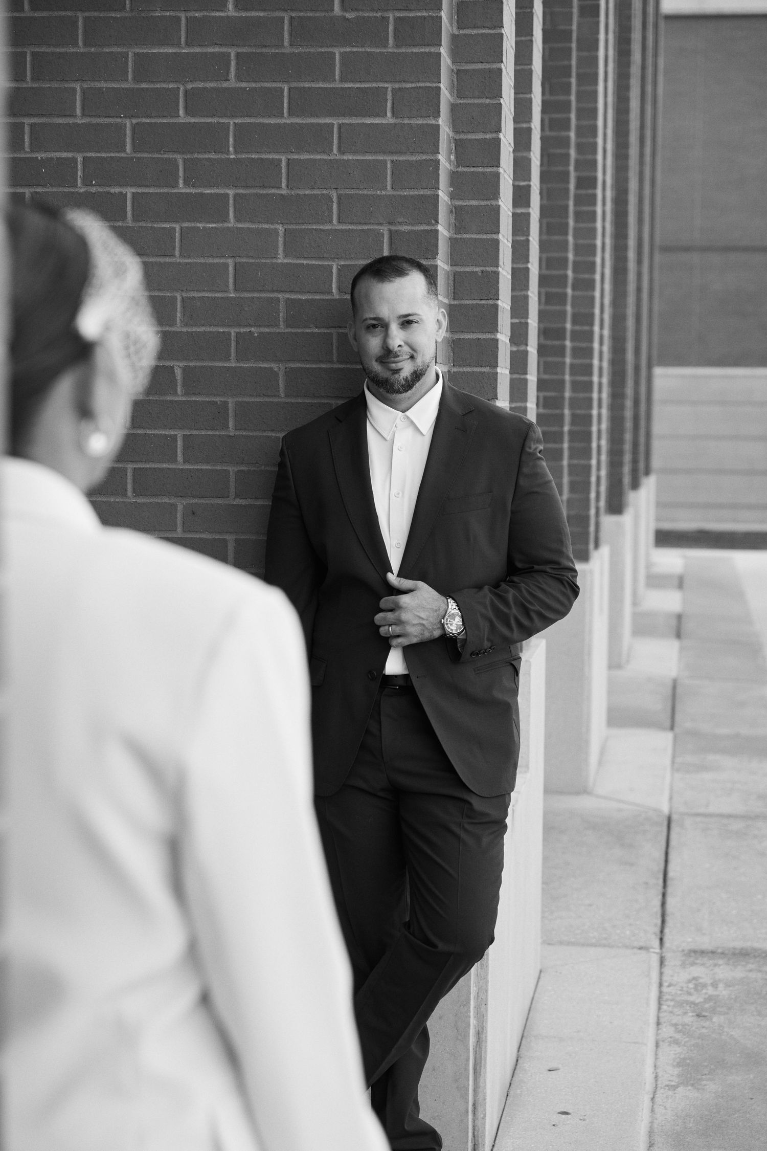 Groom in black suit leaning against brick wall smiling at bride in black and white wedding photo.