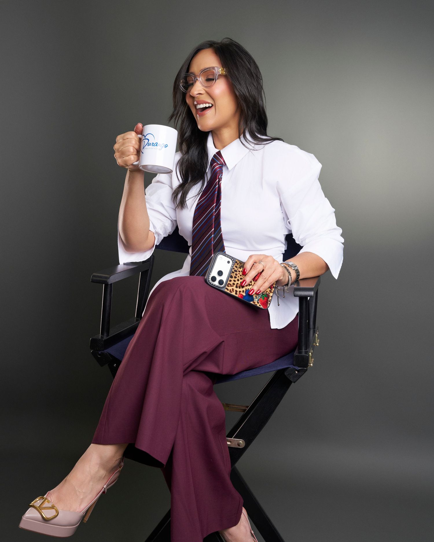 Woman in white shirt and tie sits in director chair holding coffee mug.