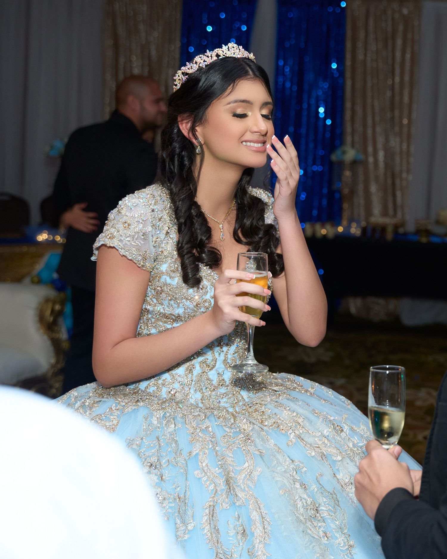 Young woman in blue and gold ball gown with tiara holds champagne glass at elegant quinceañera celebration.