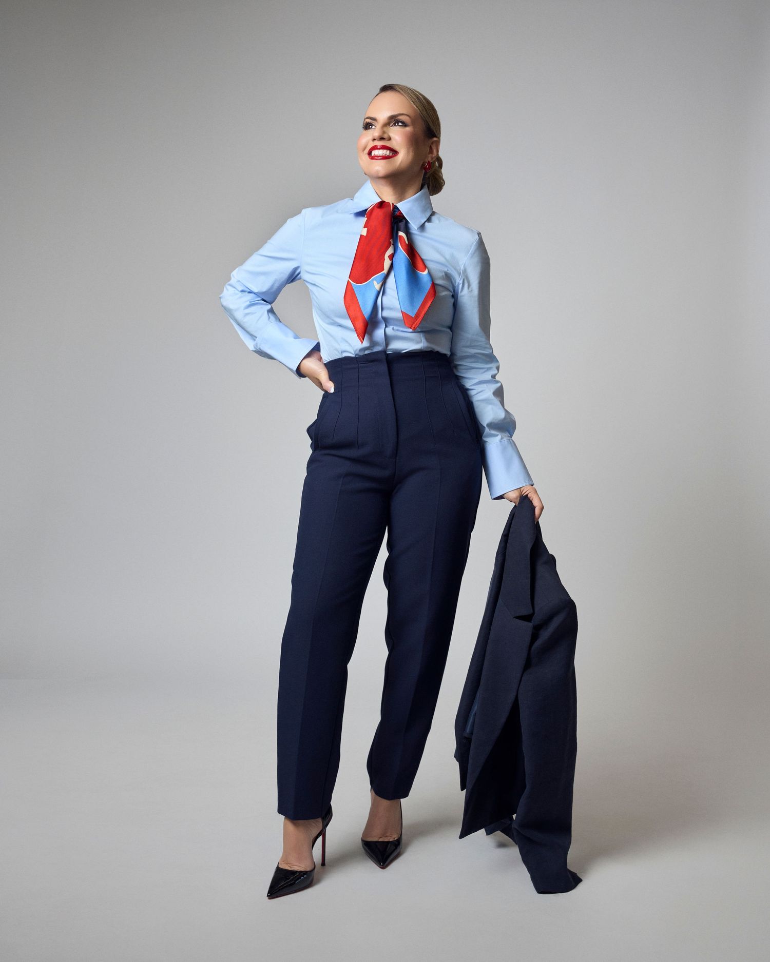 Woman in light blue shirt with red scarf and navy trousers holding a blazer on gray background.