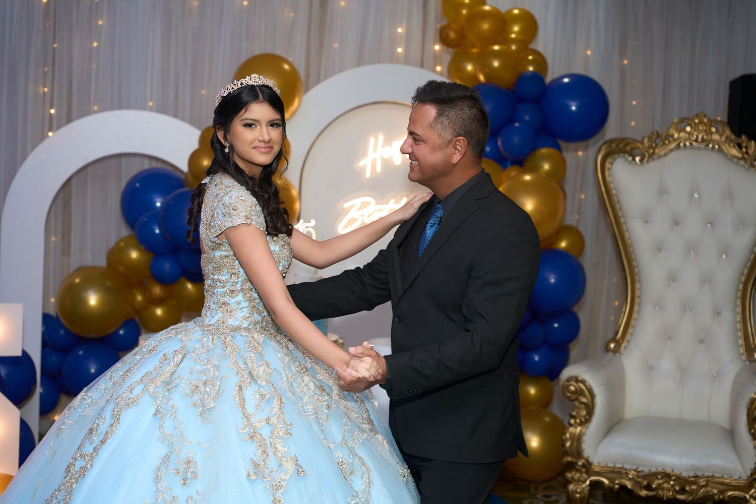 A young woman in a blue quinceañera gown dances with a man at an elegant party with gold and blue balloons.