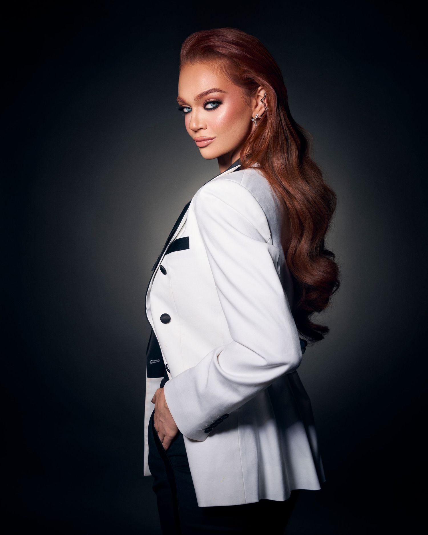 Side profile studio portrait of a woman with long auburn waves wearing a white and black blazer on dark background.