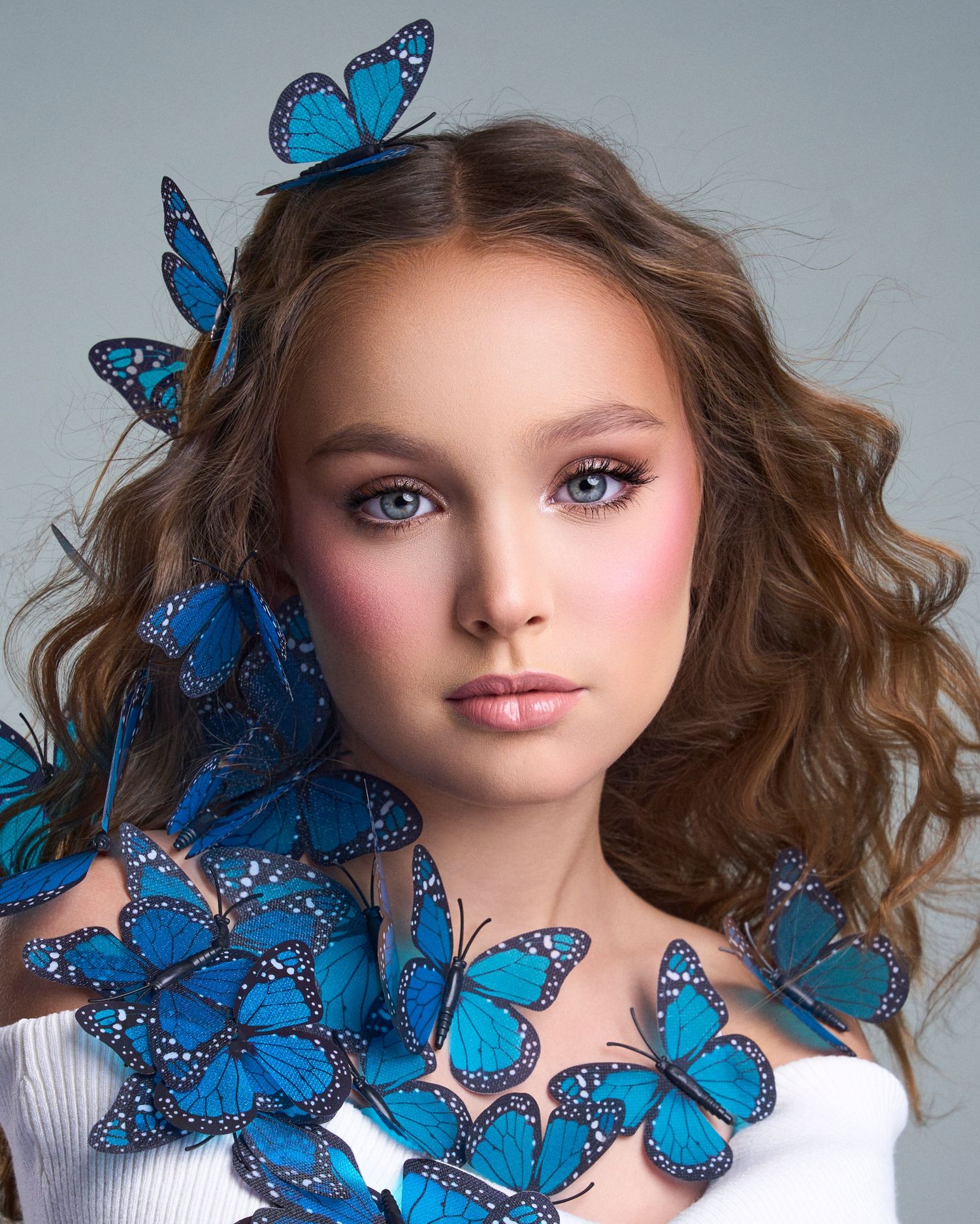 Young woman with blue eyes and rosy makeup surrounded by vibrant blue morpho butterflies in her hair and on her shoulder.