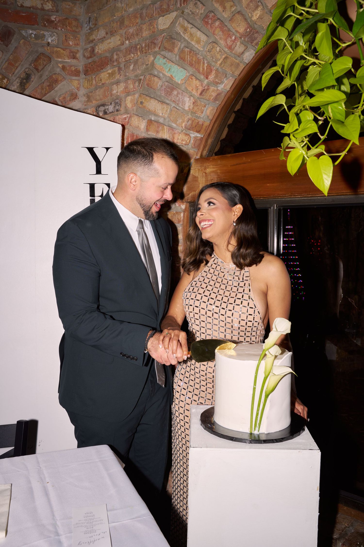 Couple smiling and cutting a white wedding cake at an elegant indoor venue with brick arched ceiling.