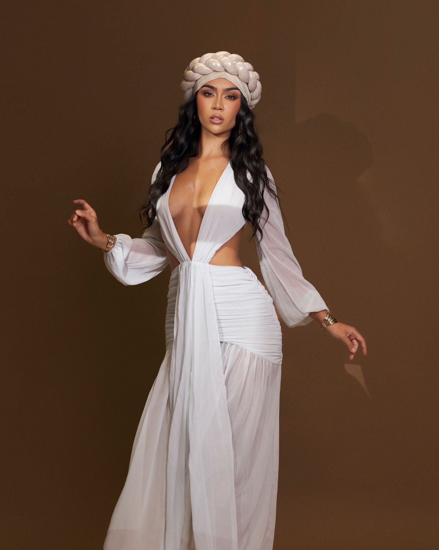 Woman in white sheer flowing dress with braided updo headpiece posing against brown background.
