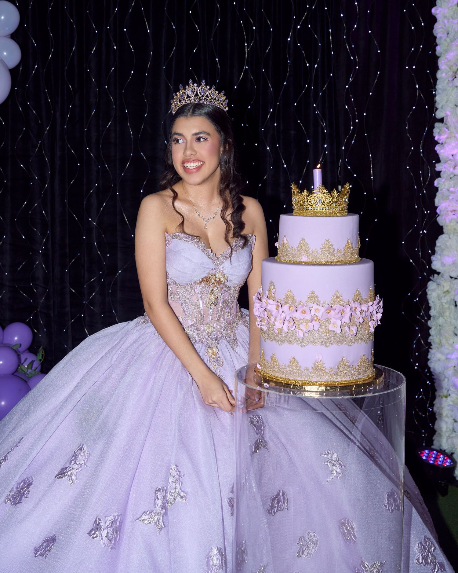 Quinceañera girl in lavender gown and crown posing beside elegant three-tier gold and white birthday cake.