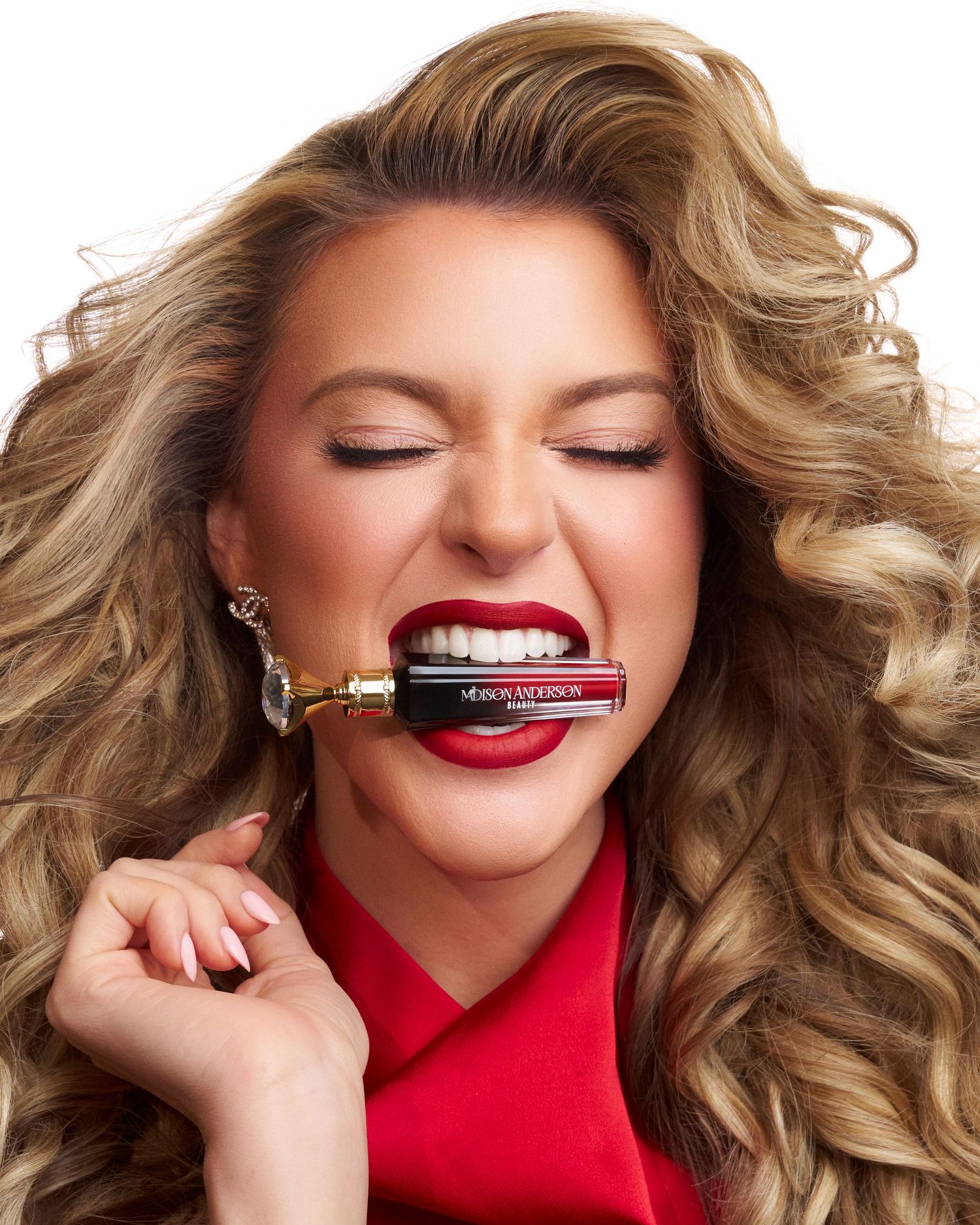 Woman with curly blonde hair biting a red lipstick tube, wearing red top and bold red lips.
